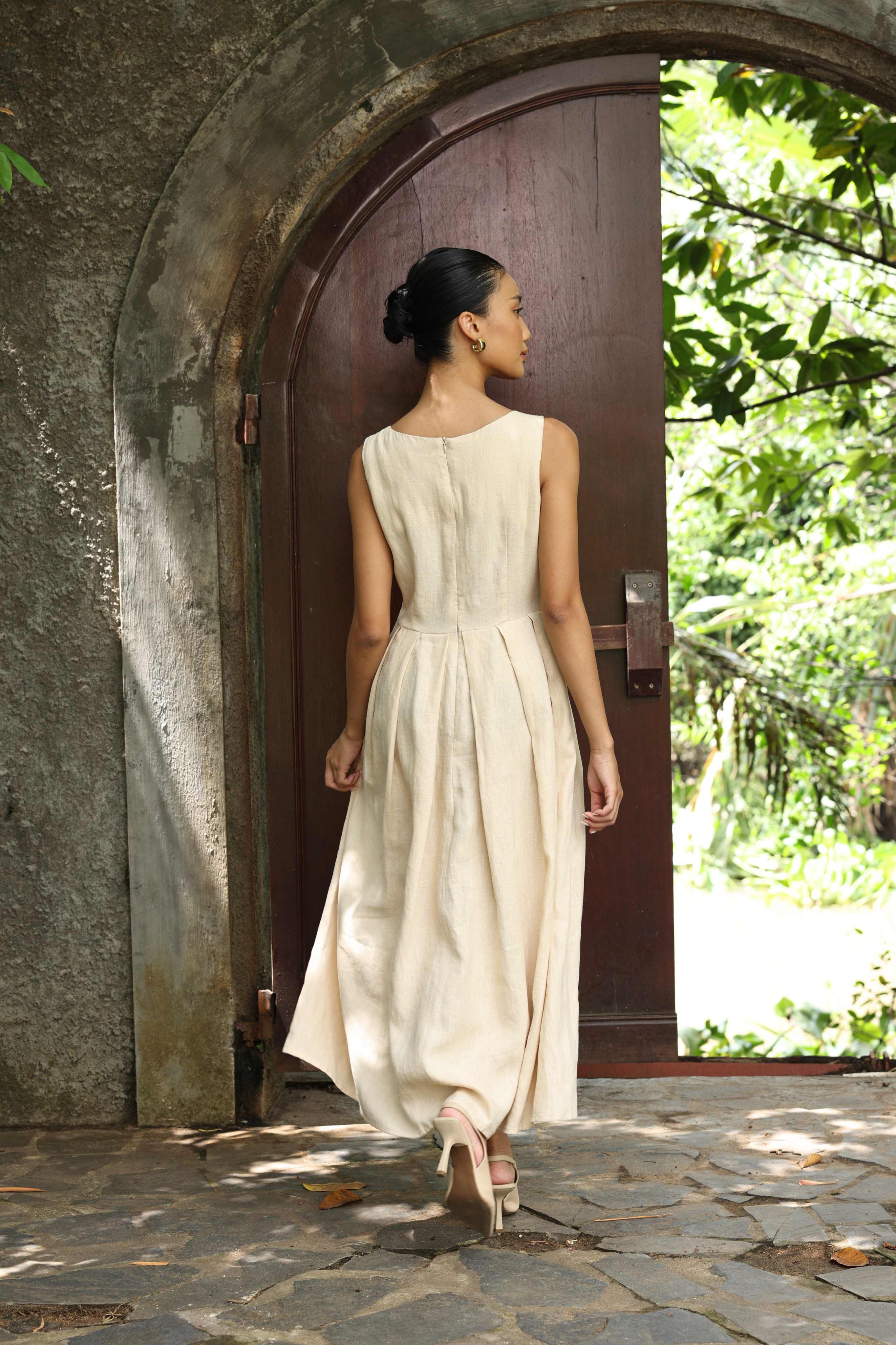 Woman in a dress standing in front of a stone archway with greenery outside.