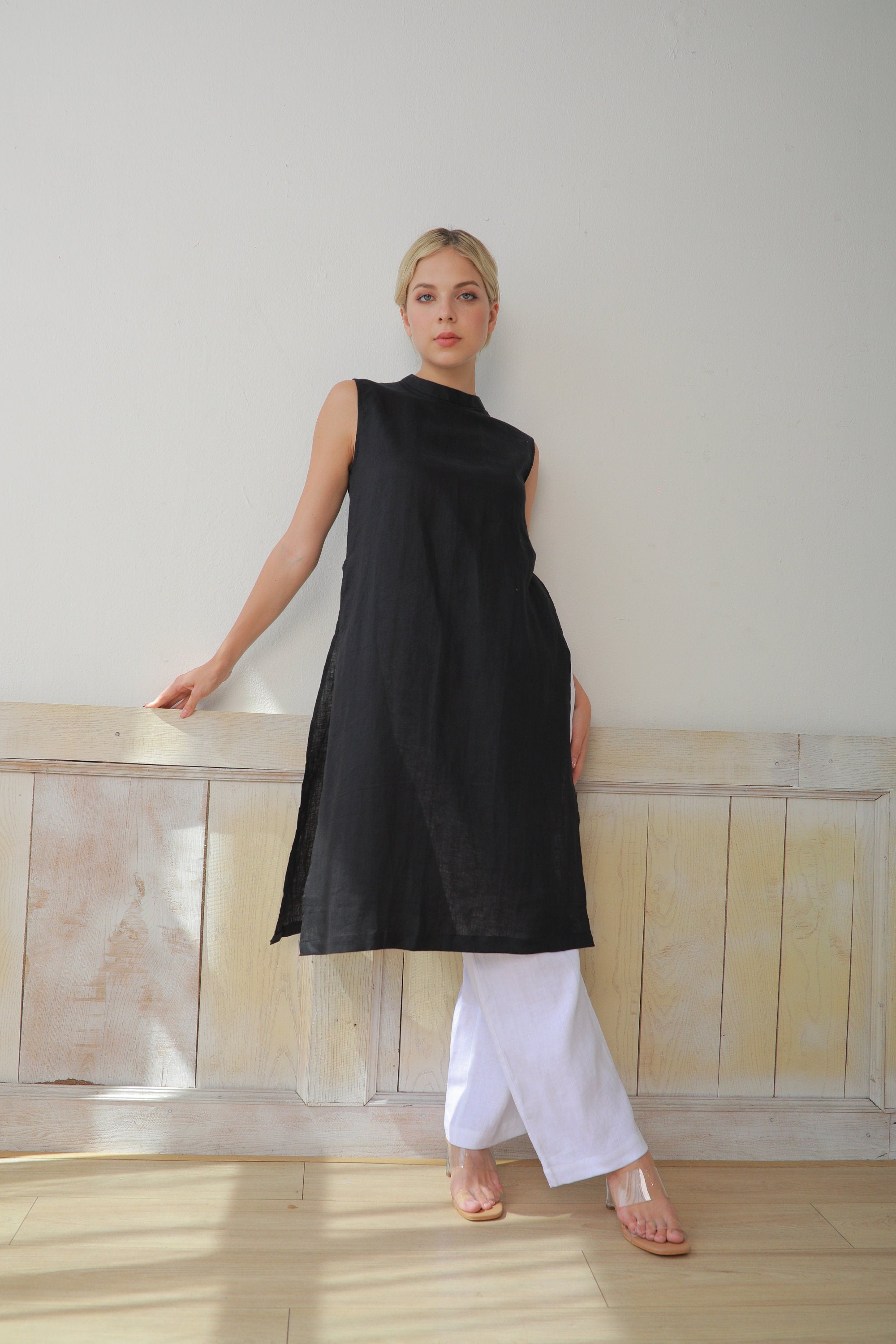 Woman wearing a black sleeveless dress and white pants against a light-colored wall.