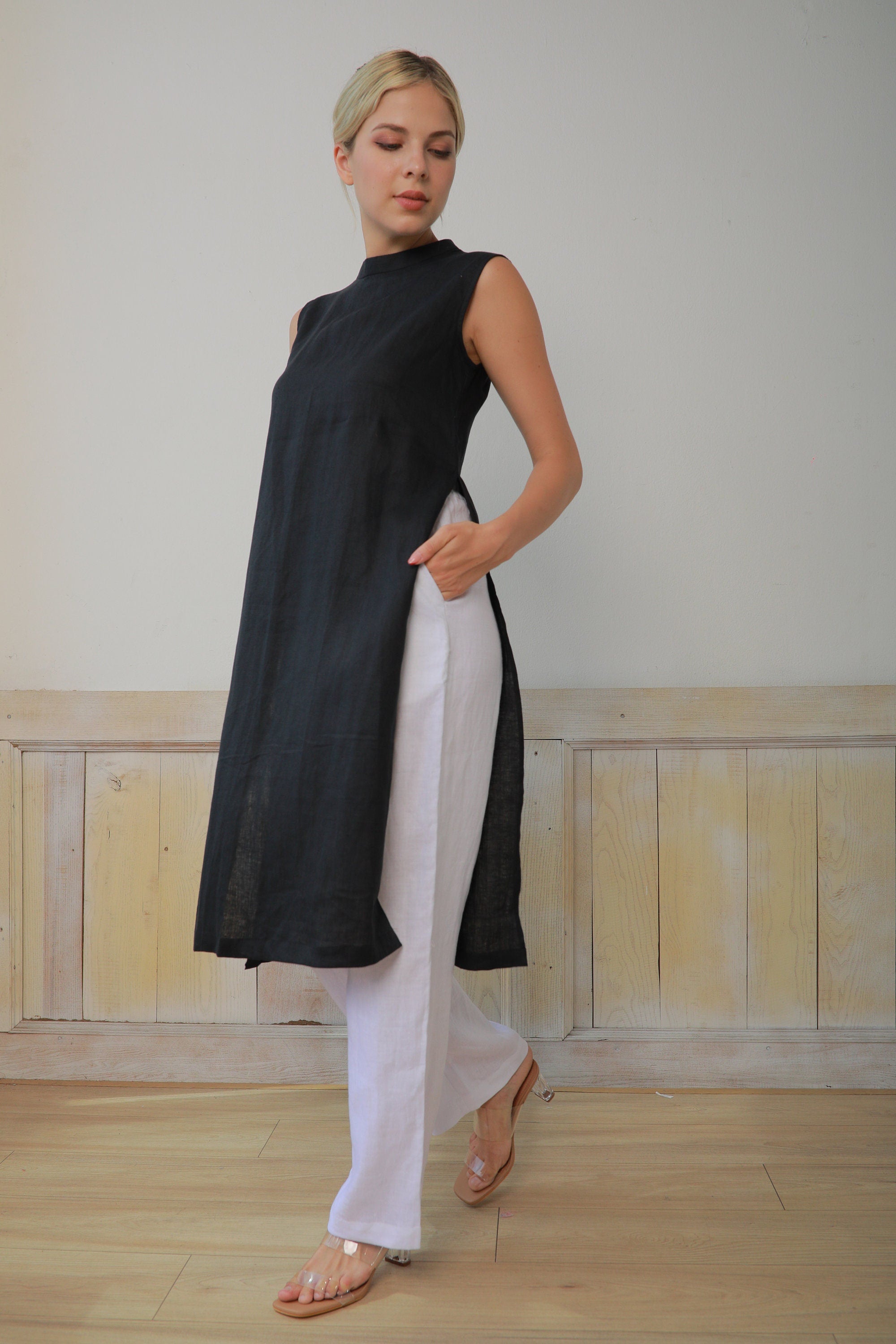 Woman wearing a black sleeveless top and white pants in a minimalistic room.