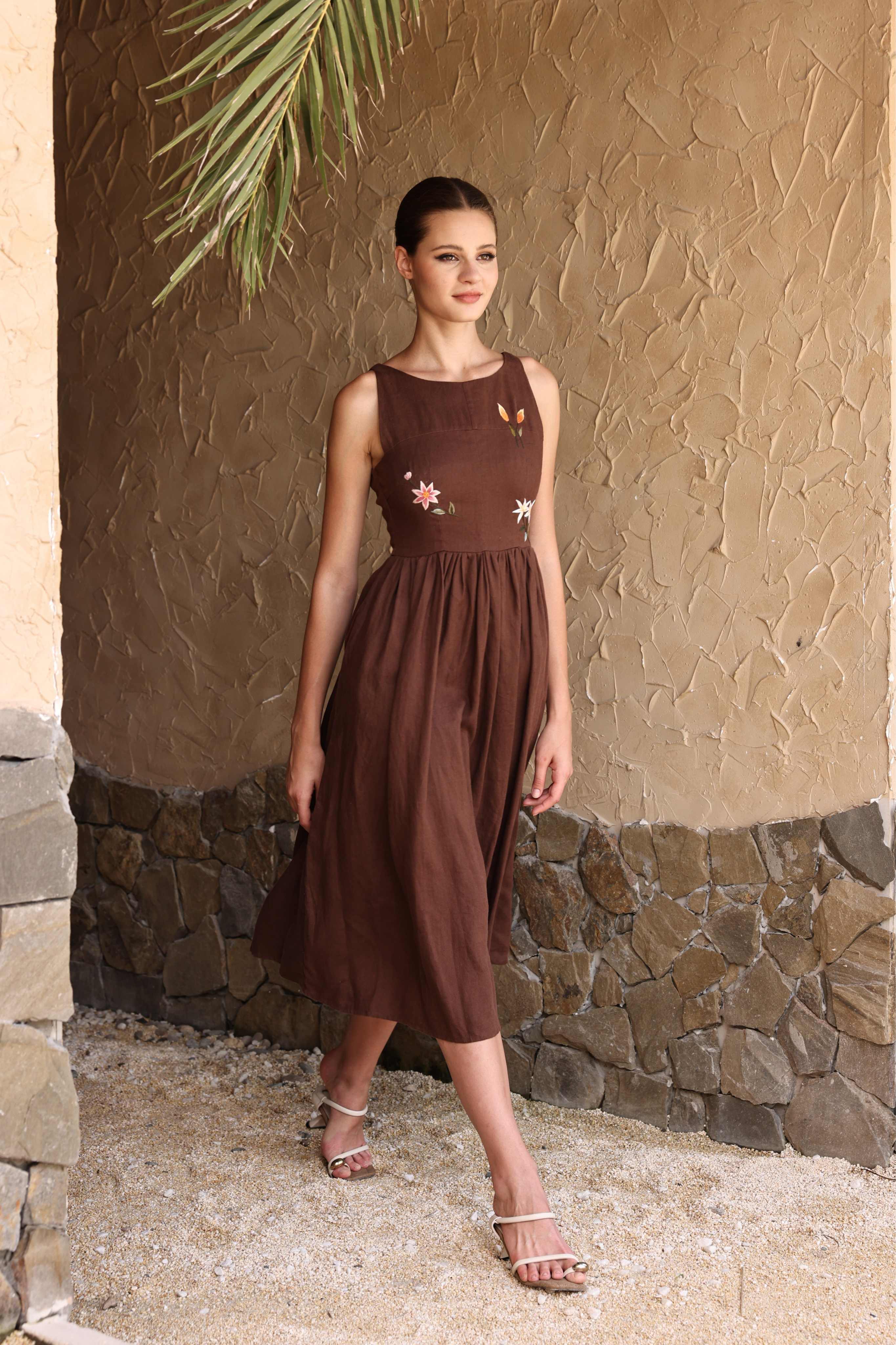 Woman wearing a brown dress with floral embellishments against a stone wall.