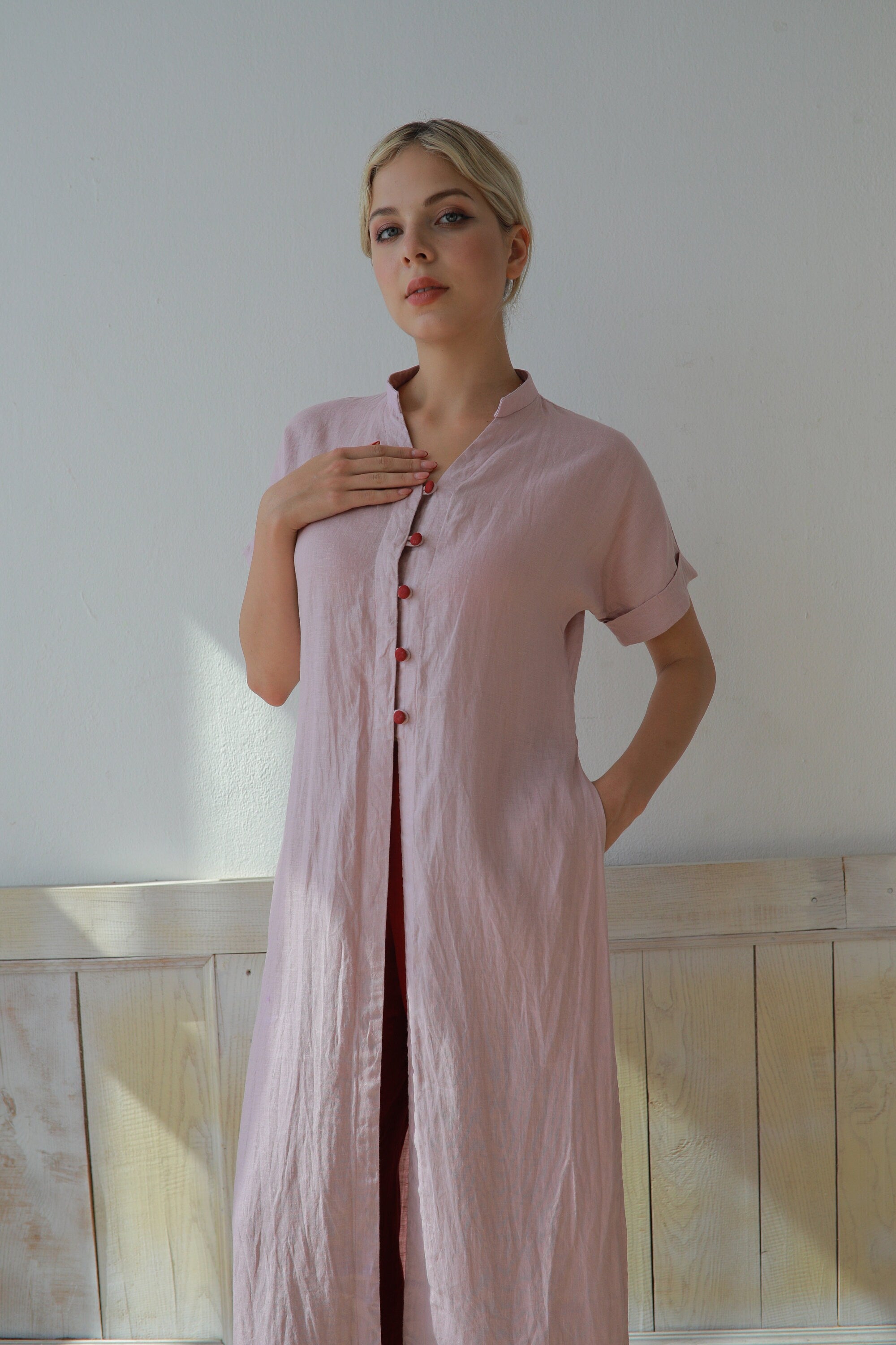 Woman wearing a long pink dress with buttons against a white wall.