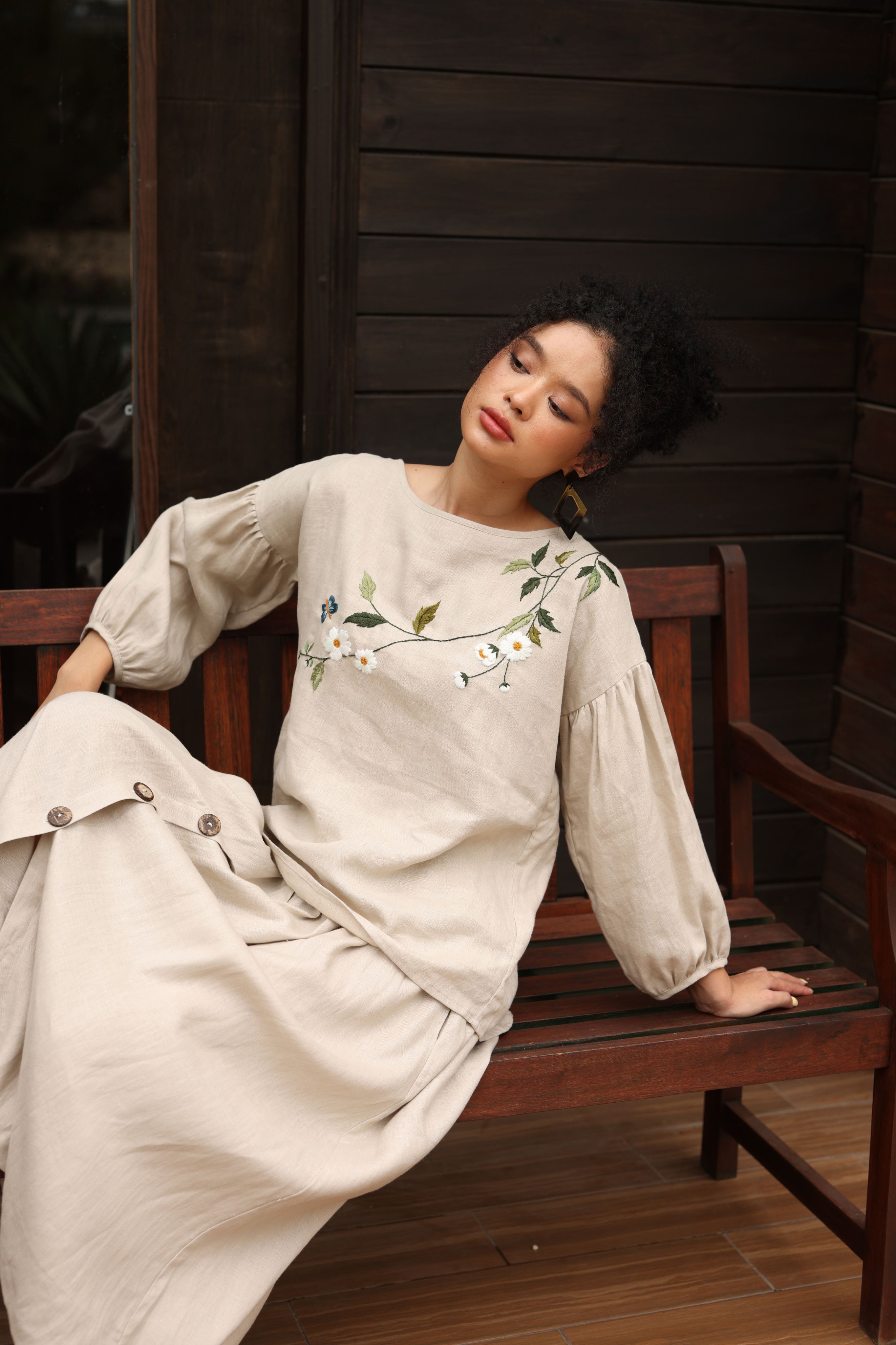 Woman in a beige dress with floral embroidery sitting on a wooden bench.