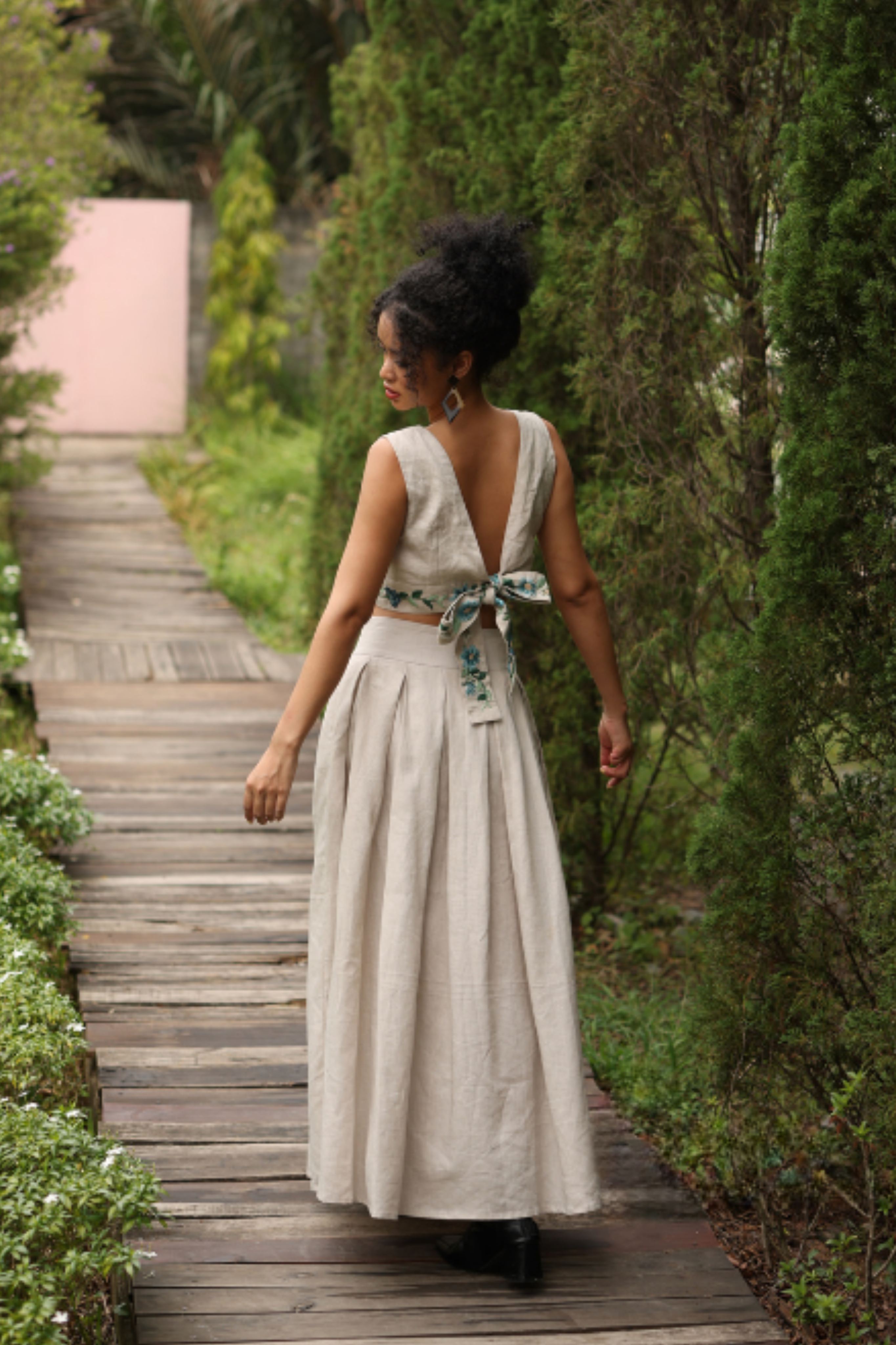 Woman in a light-colored outfit walking on a wooden path through a garden.
