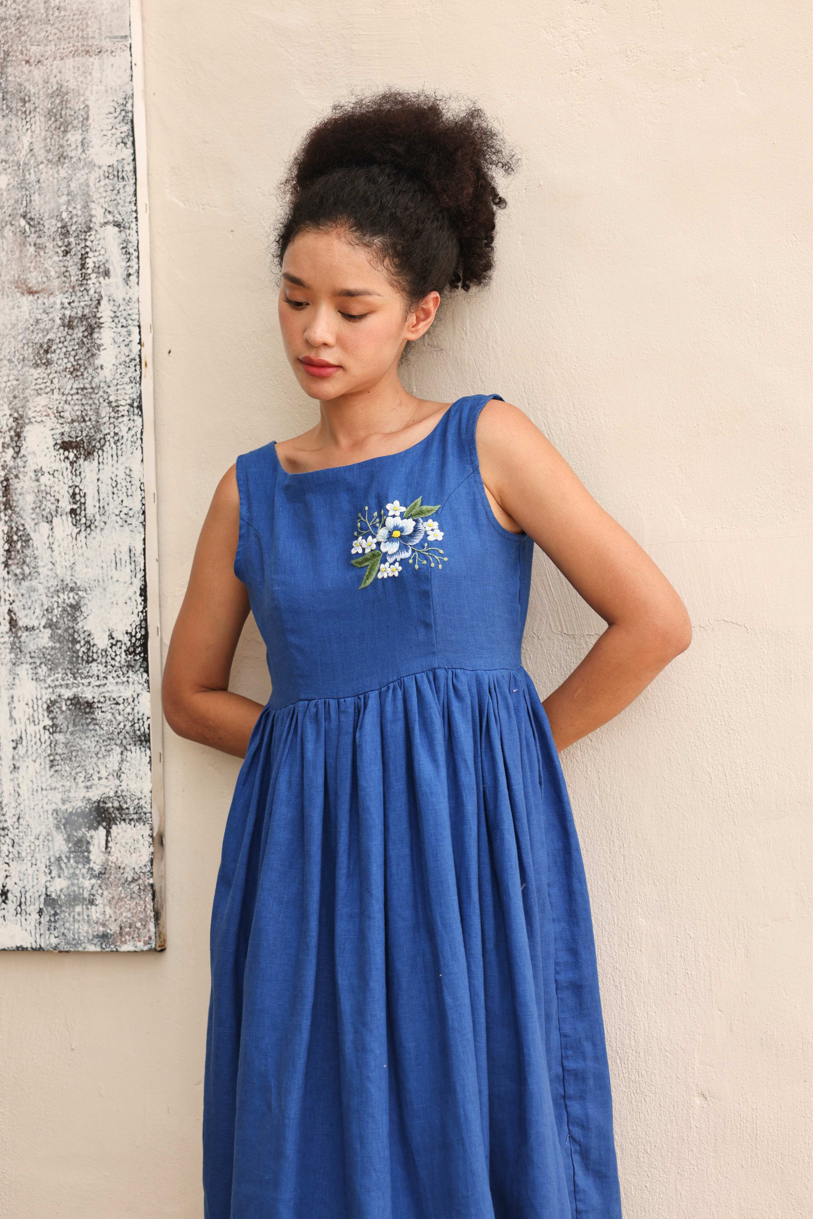 Woman wearing a blue dress with floral embroidery against a neutral wall.