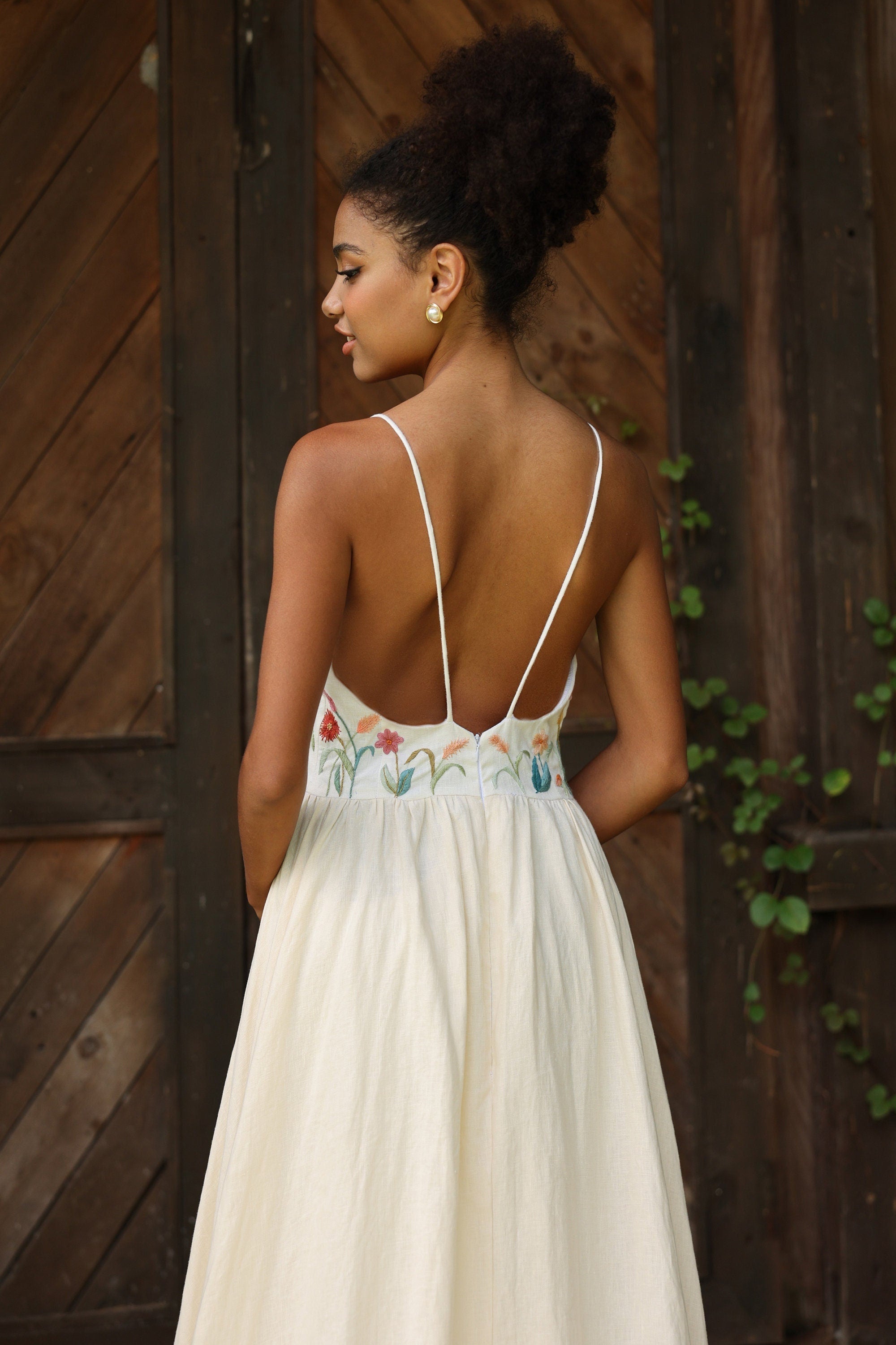 Woman wearing an ivory dress with floral embroidery in front of a wooden wall.