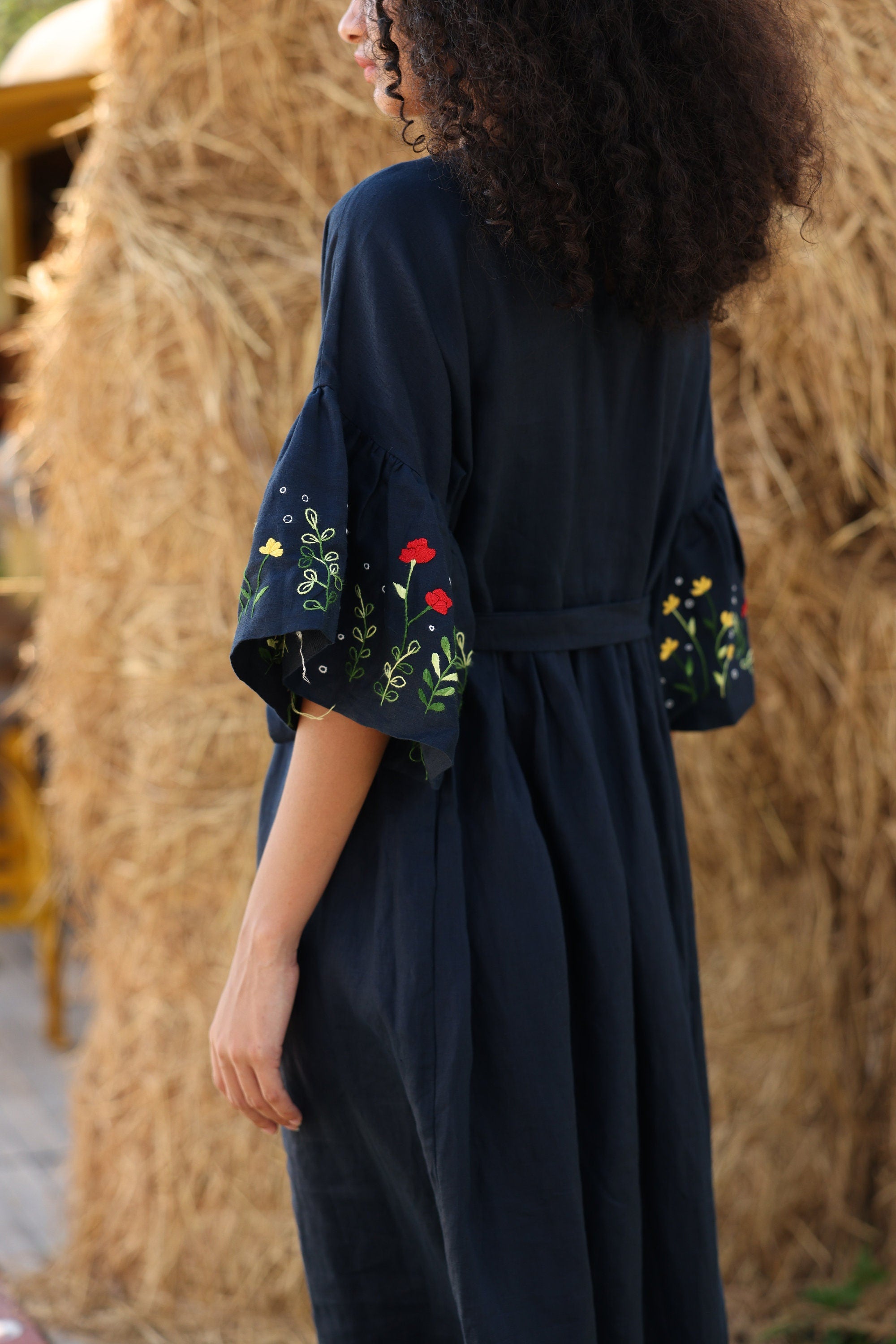 Person wearing a dark blue dress with floral embroidery in front of hay bales.