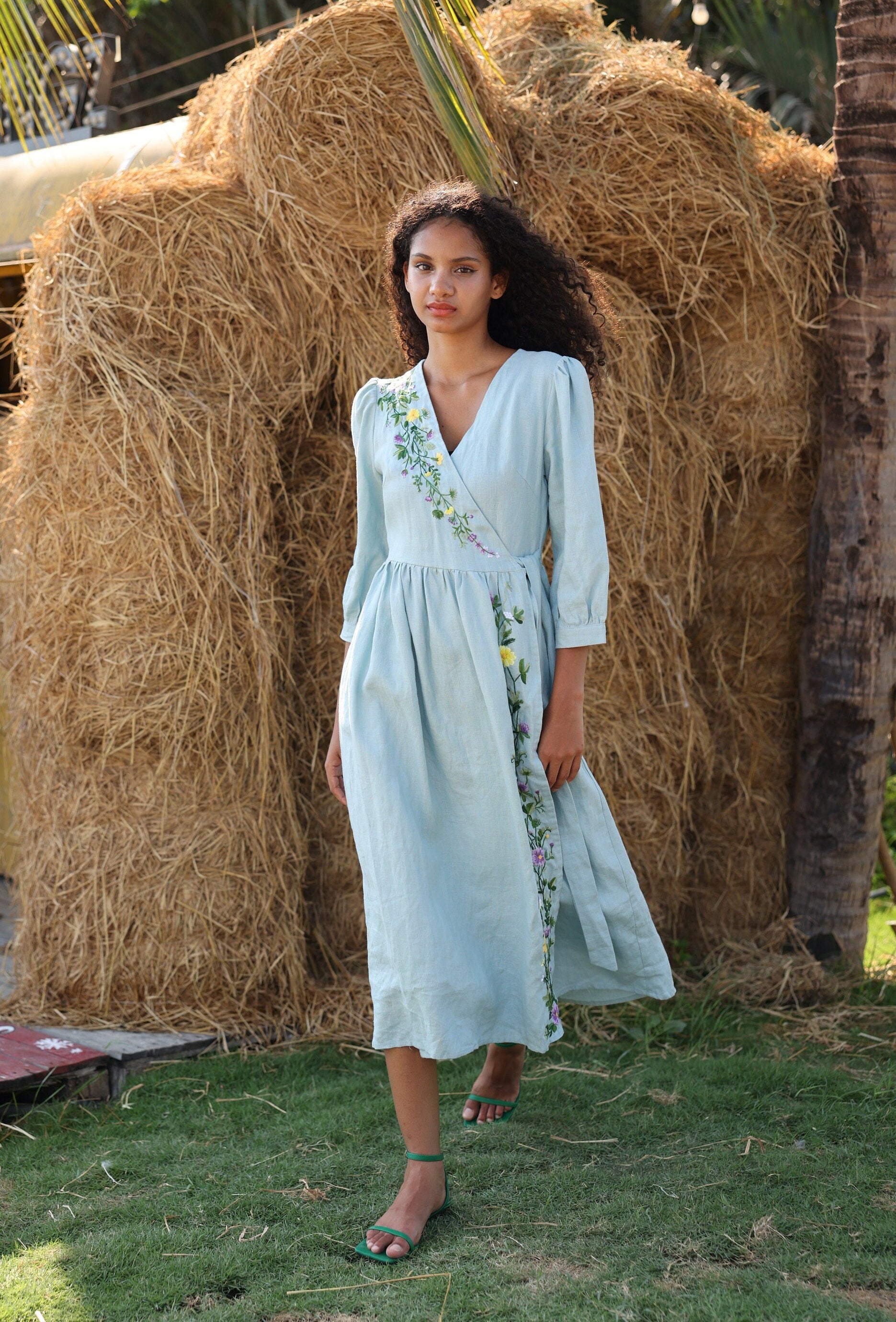 Woman in a light blue dress standing in front of a large hay bale.