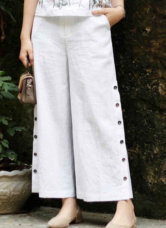 Person wearing white pants with buttons against a dark wall.