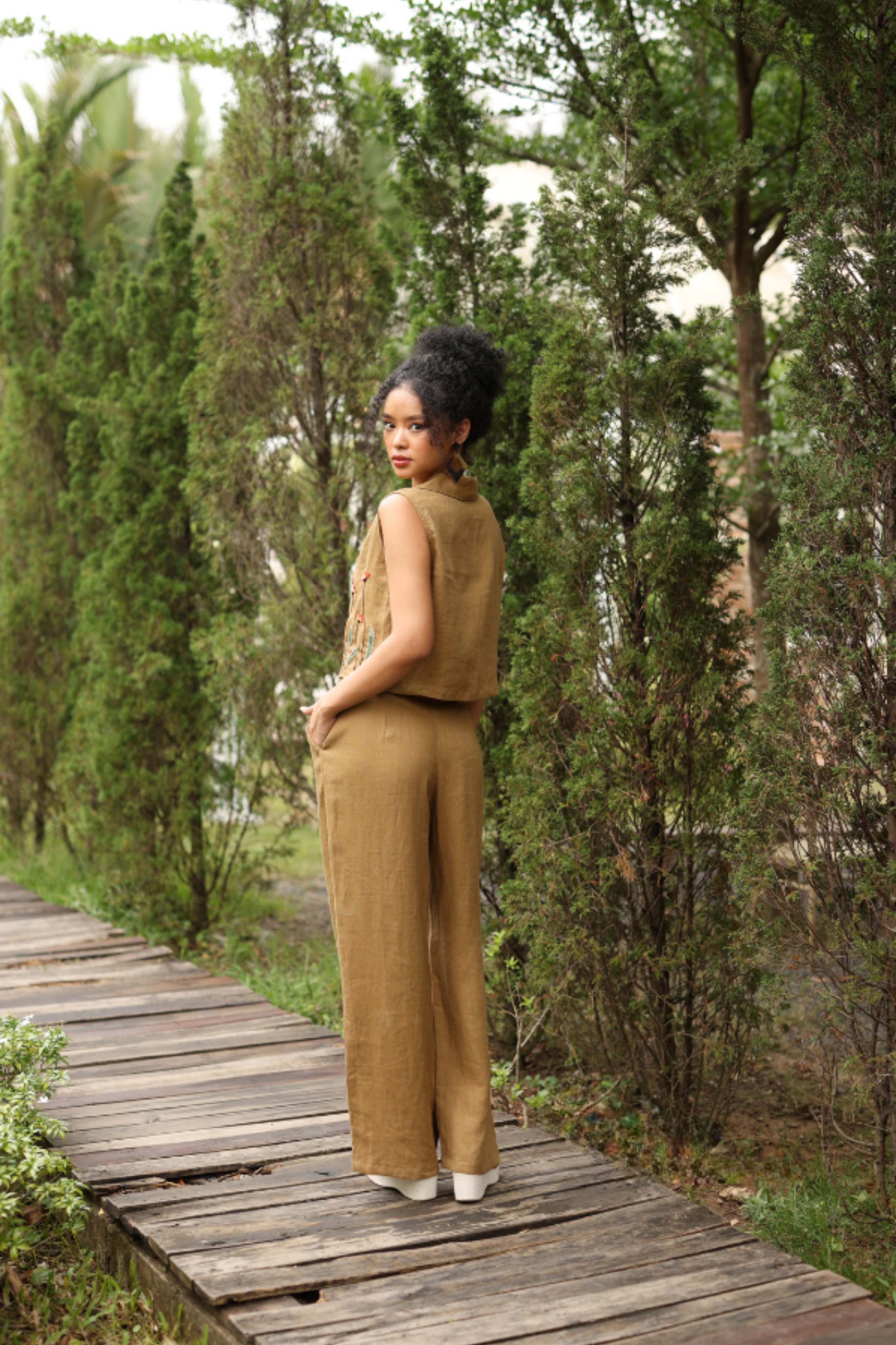 Woman in a brown outfit standing on a wooden path surrounded by greenery