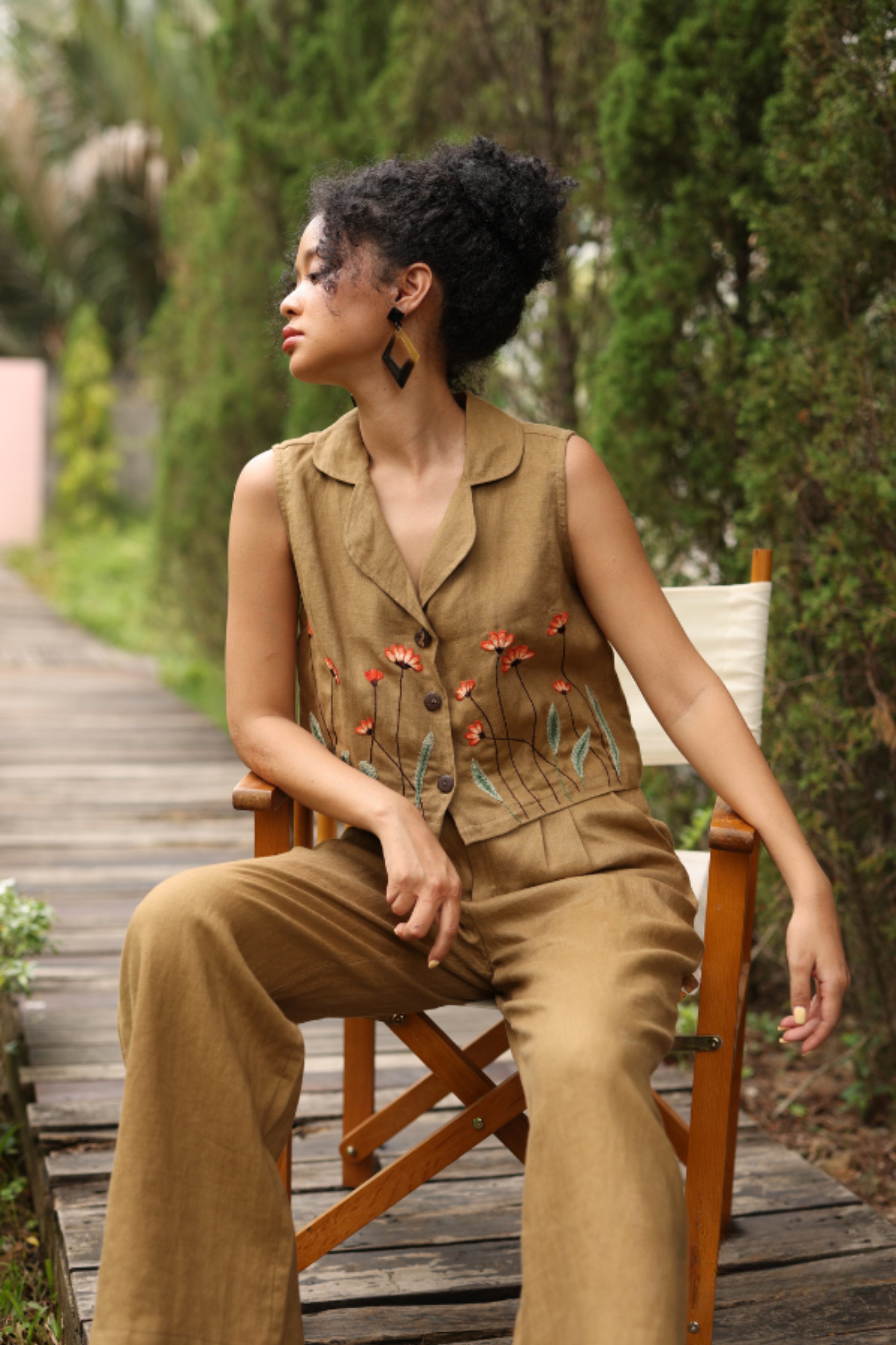 Woman wearing an embroidered vest and pants sitting on a wooden chair outdoors.