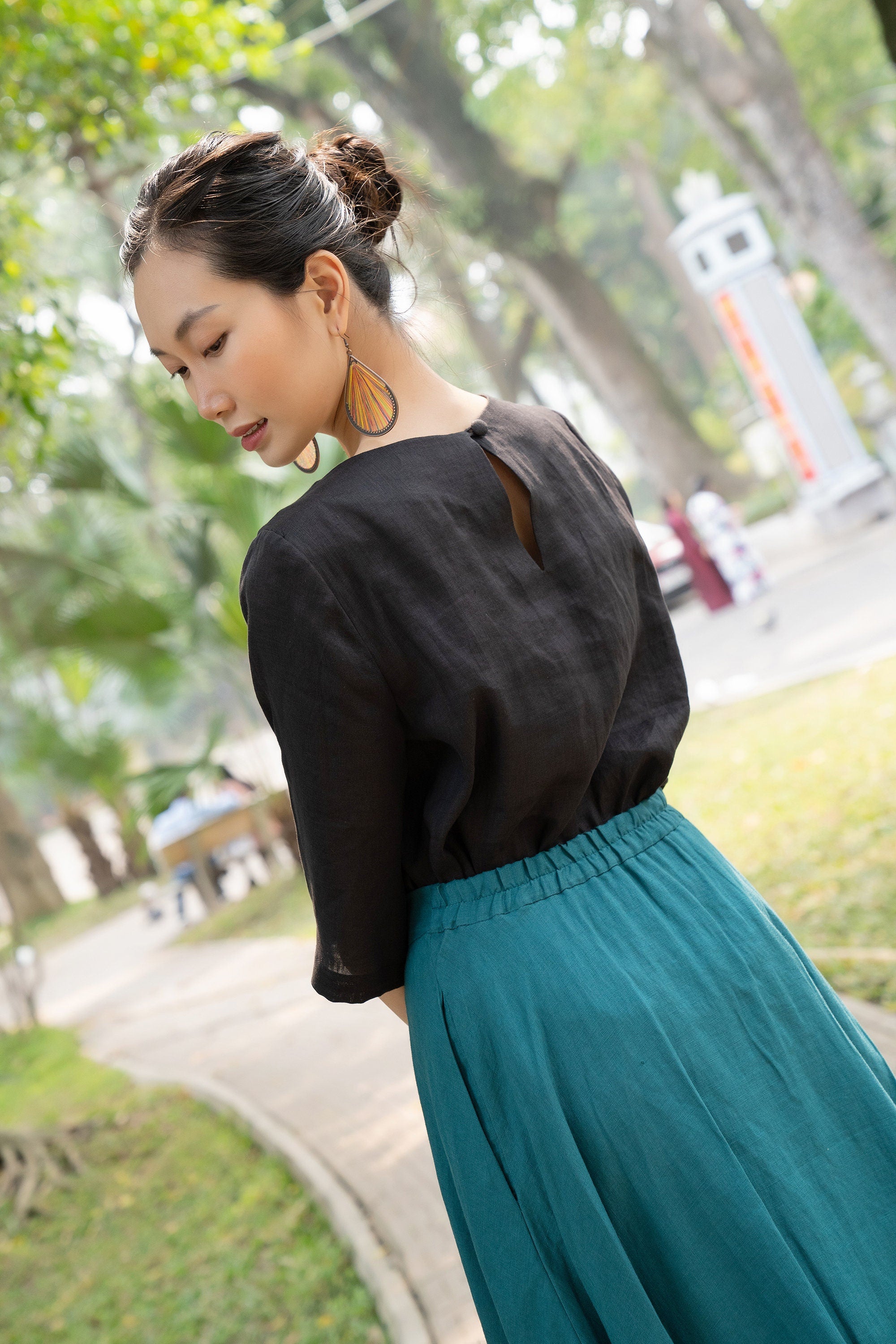 Woman in black top and teal skirt standing outdoors with trees and a path in the background
