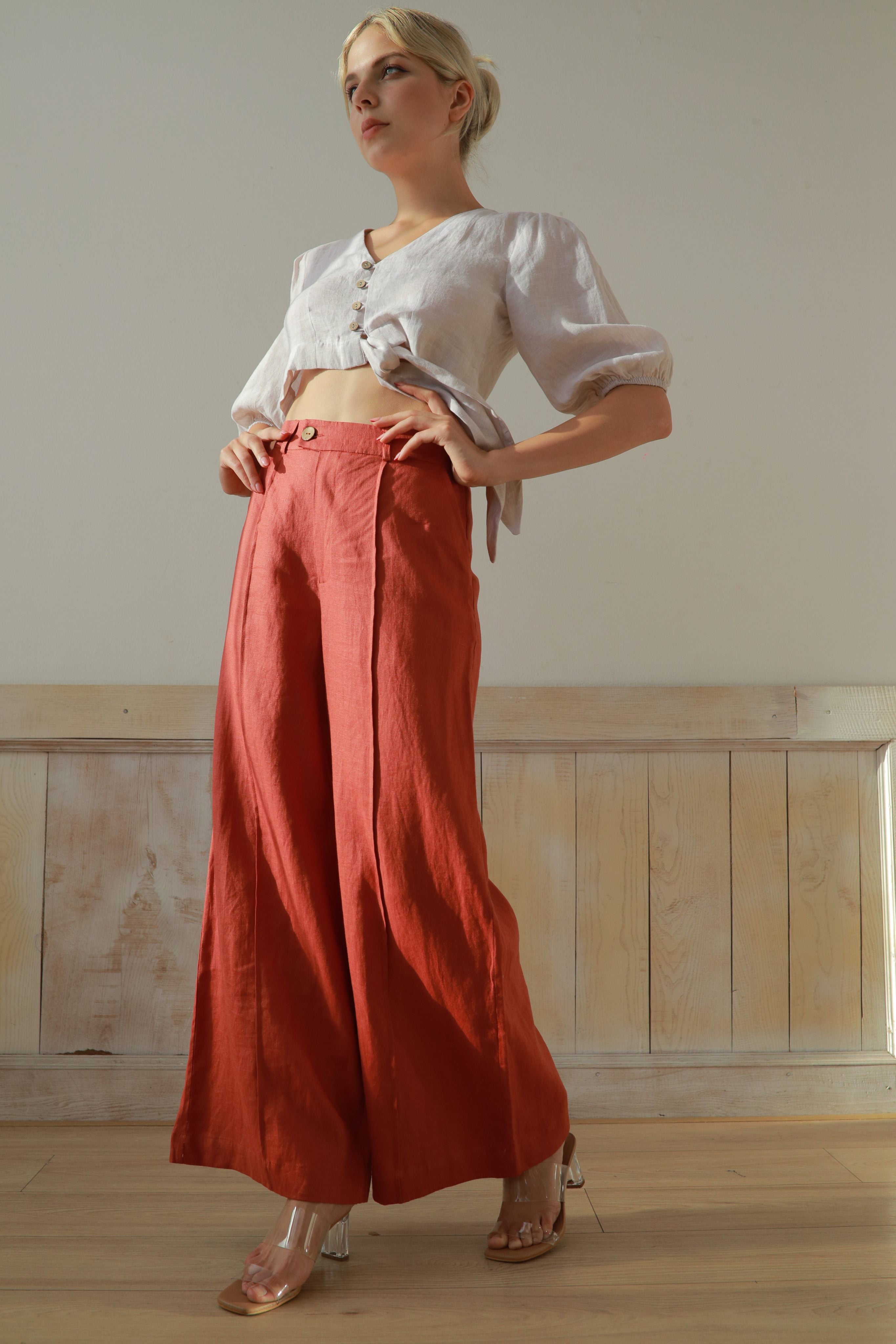 Woman wearing a white blouse and red pants standing against a neutral wall.