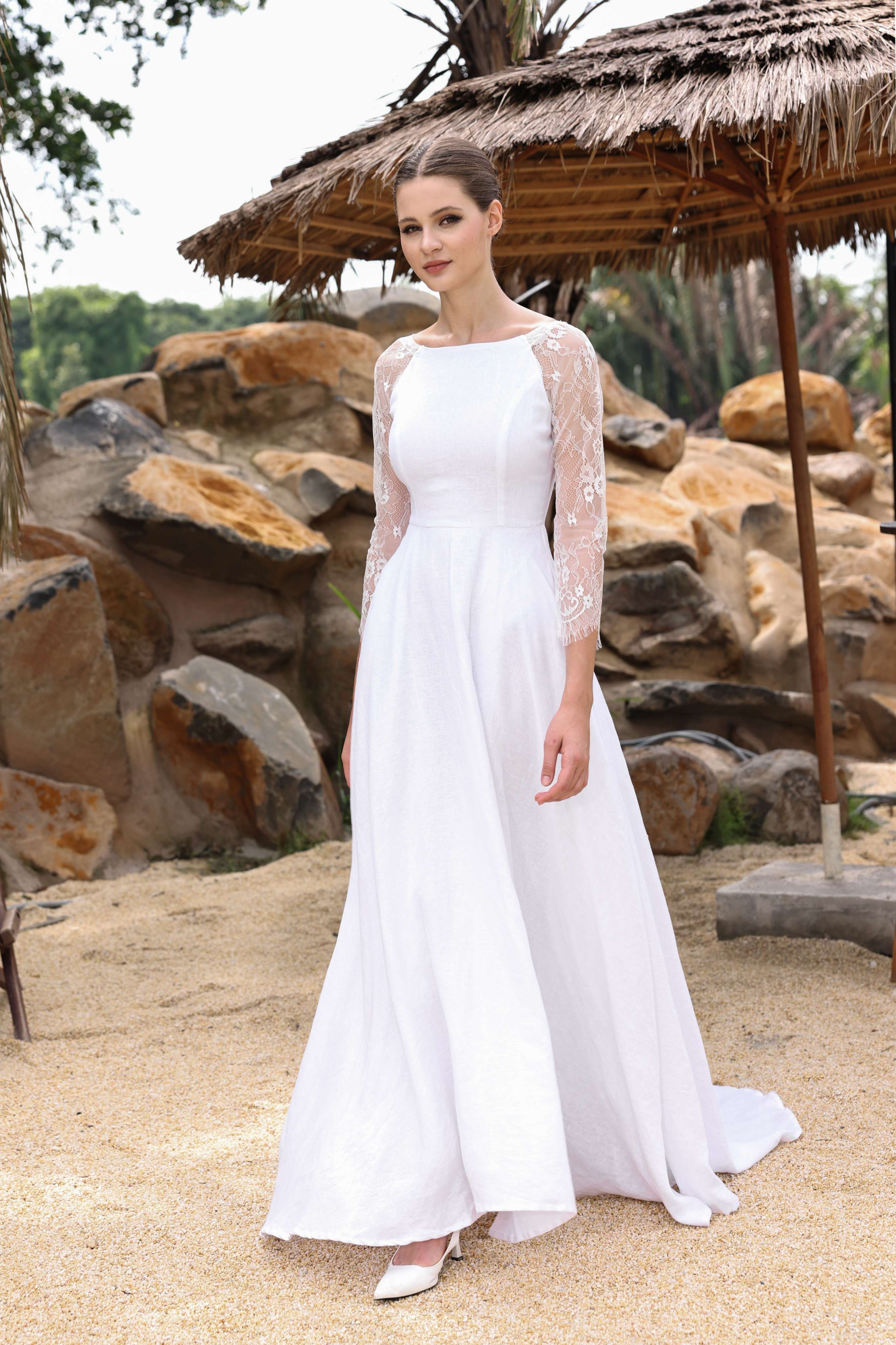 Woman in a white dress with a sheer overlay standing under a thatched umbrella with a natural rock and tree background.