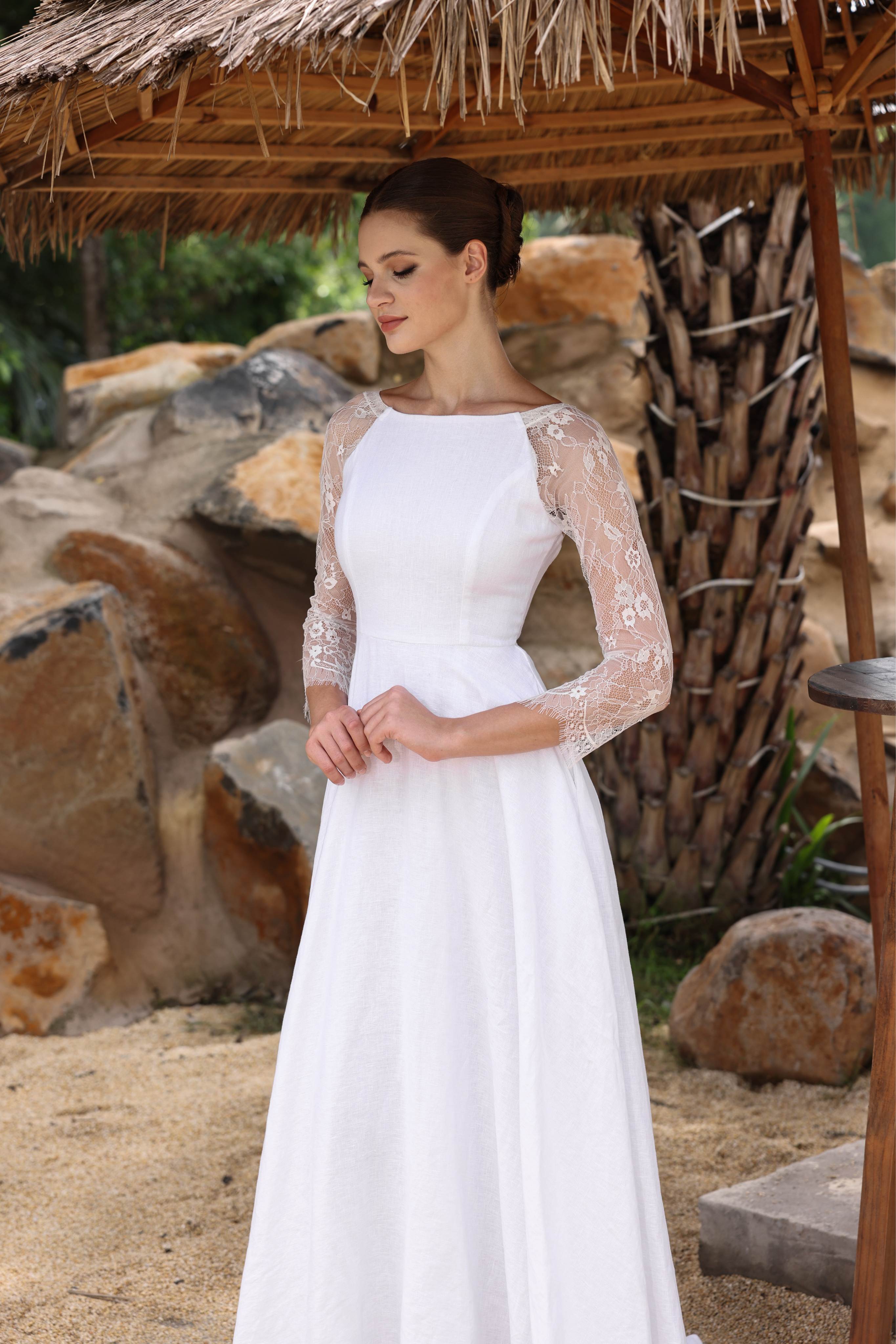 Woman in a white dress with lace sleeves standing under a thatched roof in a natural setting.