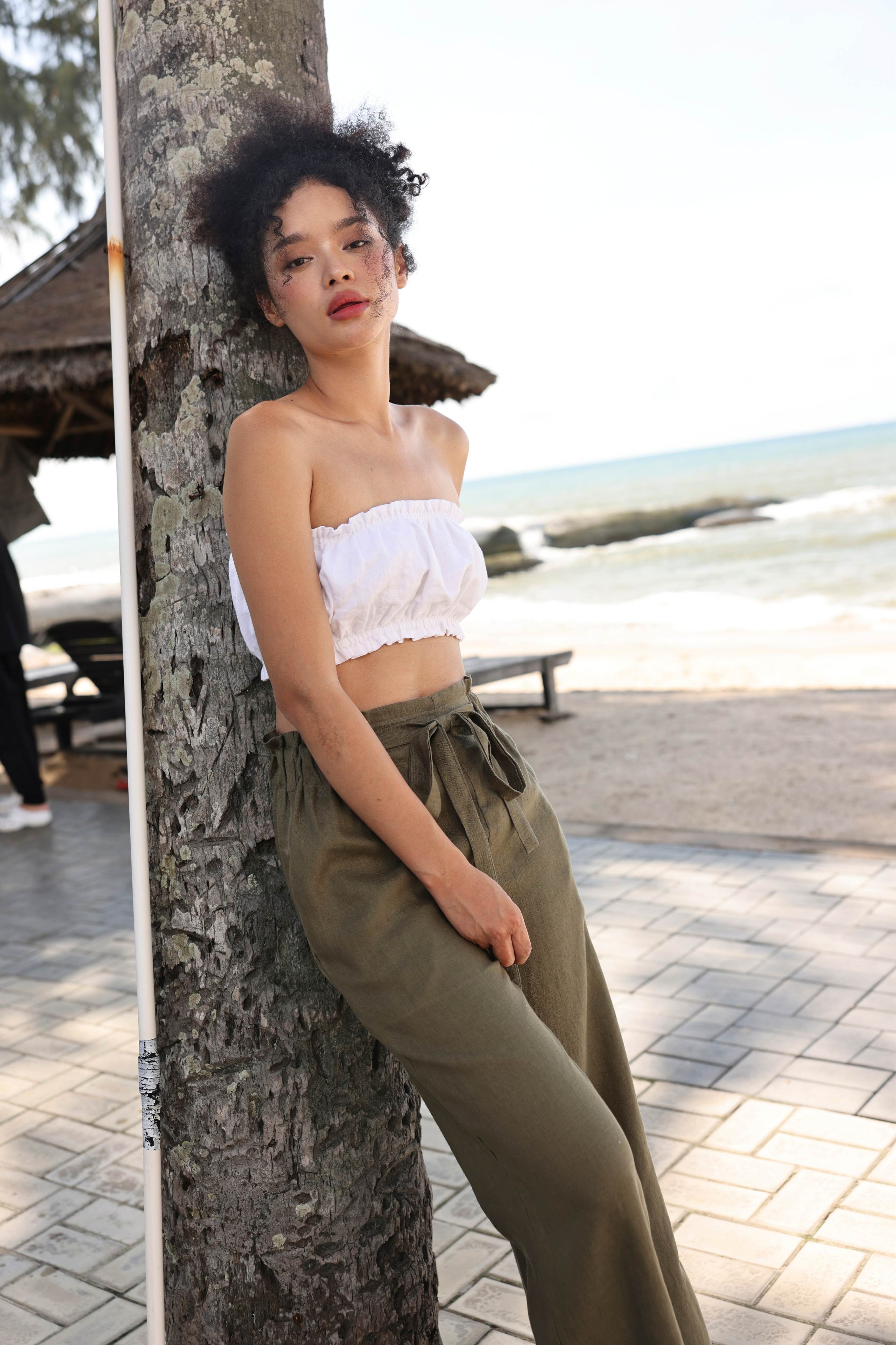 Woman in a white top and green pants leaning against a tree by a beach.