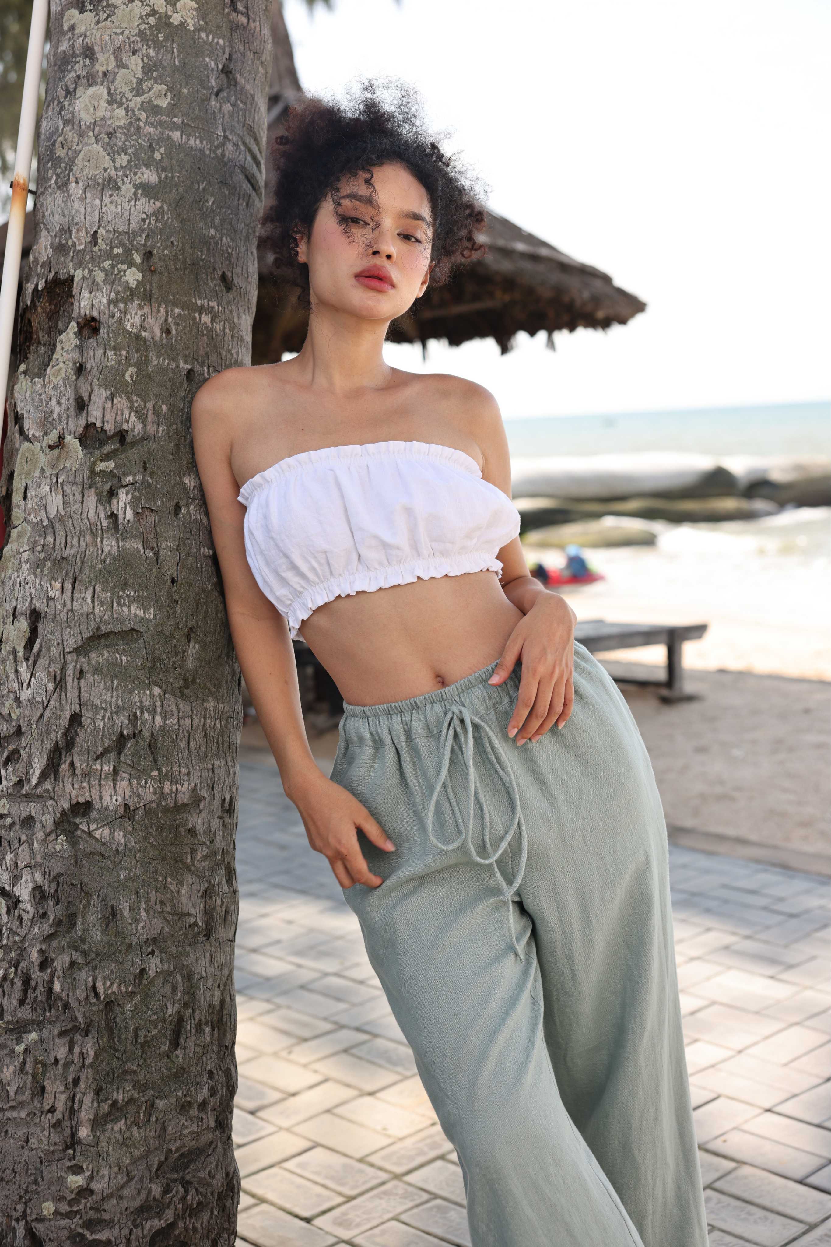 Woman in a white top and light green pants leaning against a tree by a beach.