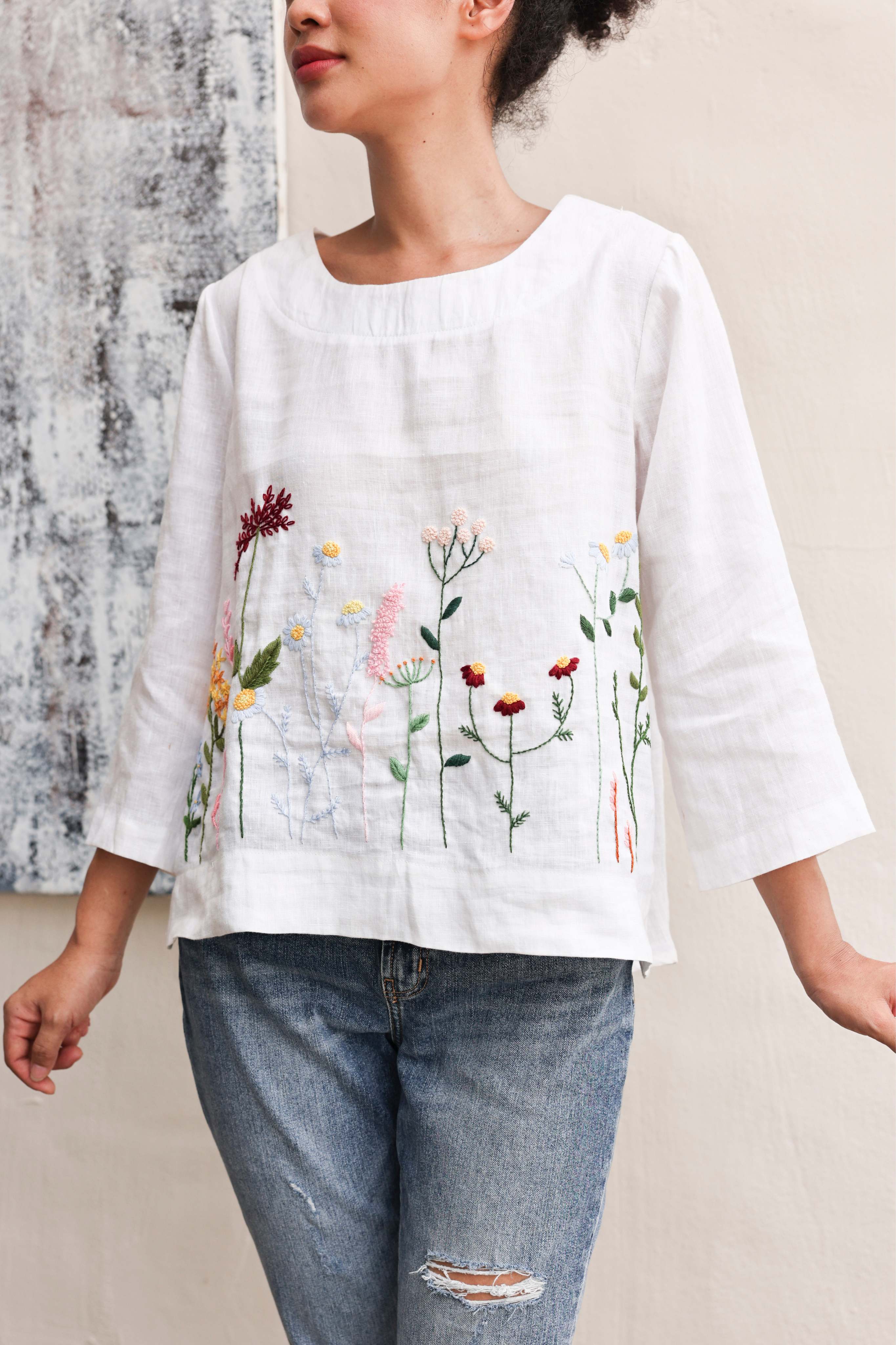 Person wearing a white blouse with floral embroidery and blue jeans against a neutral background