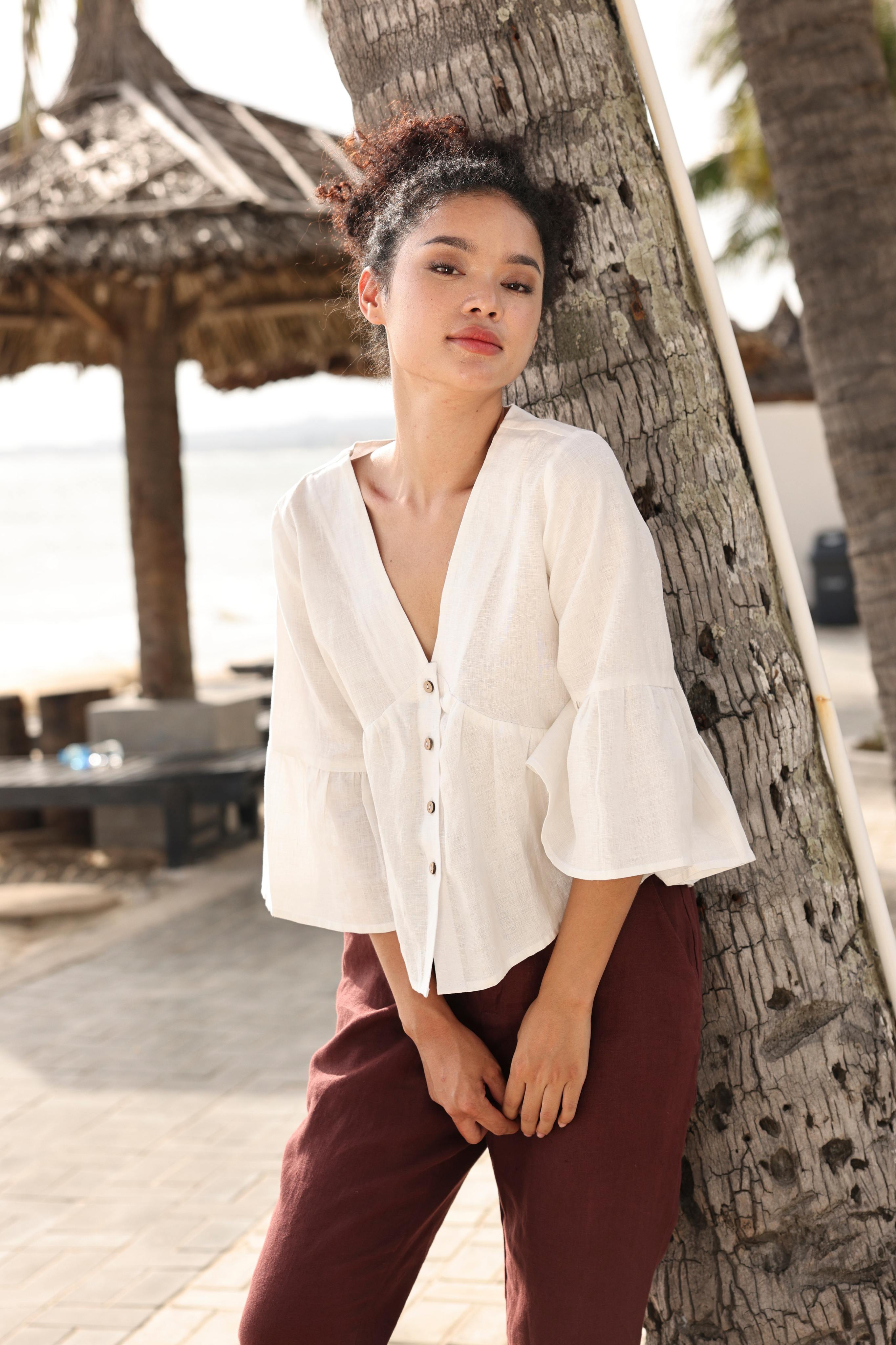 Woman in a white blouse and maroon pants leaning against a tree in a tropical setting.