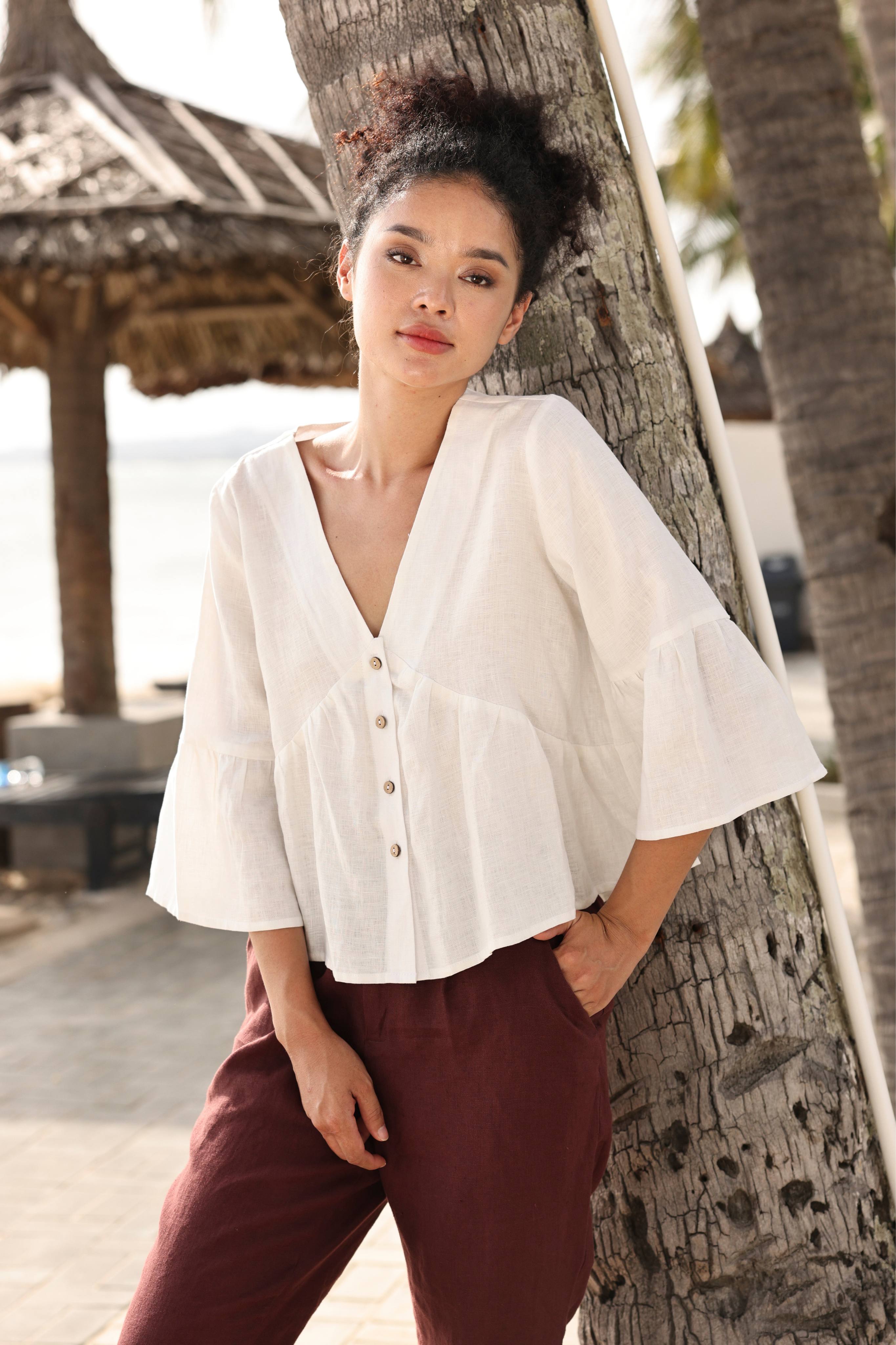 Woman in a white blouse and maroon pants leaning against a tree by a beach.