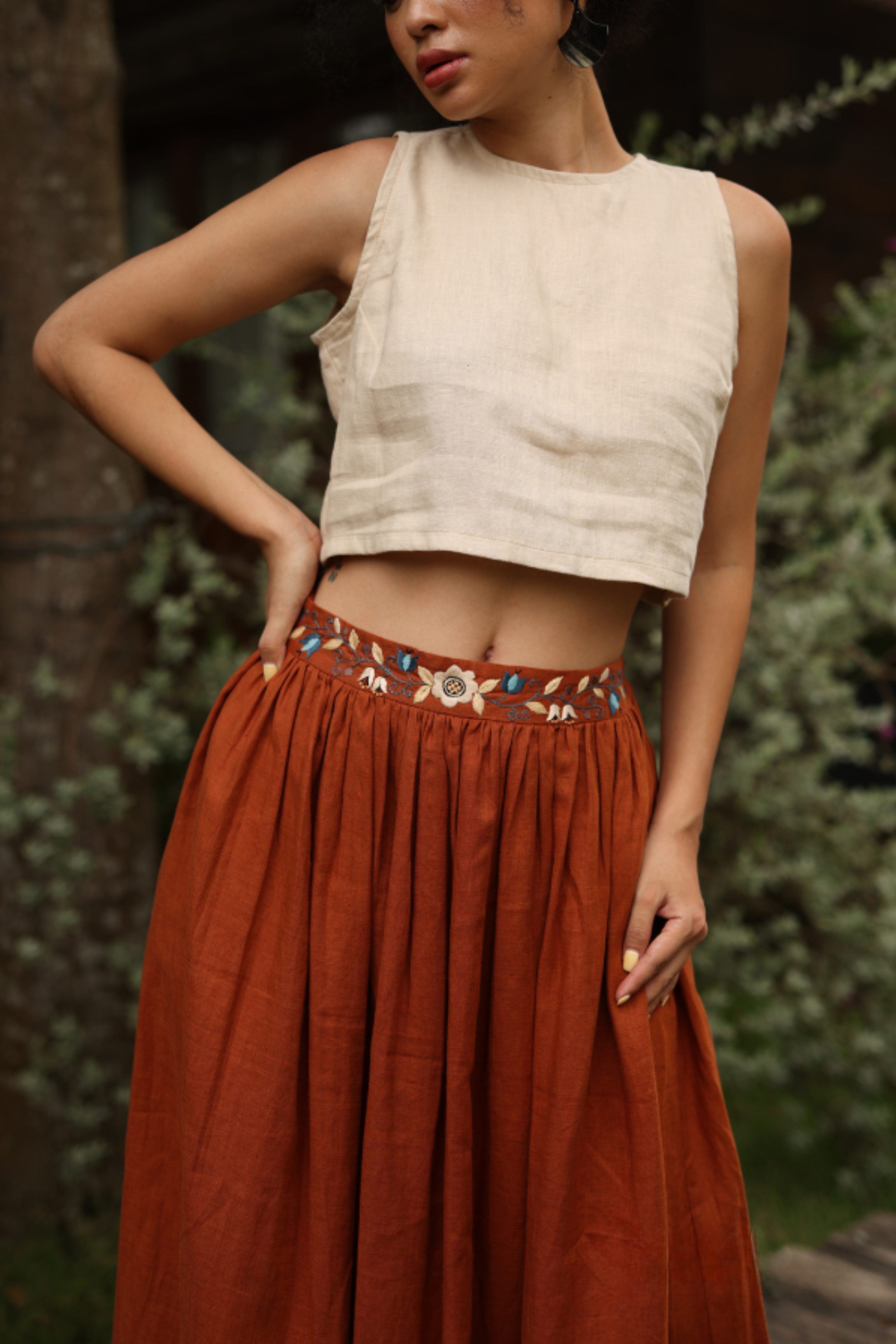 Woman wearing a beige top and rust-colored skirt with embroidery, standing against a natural background.