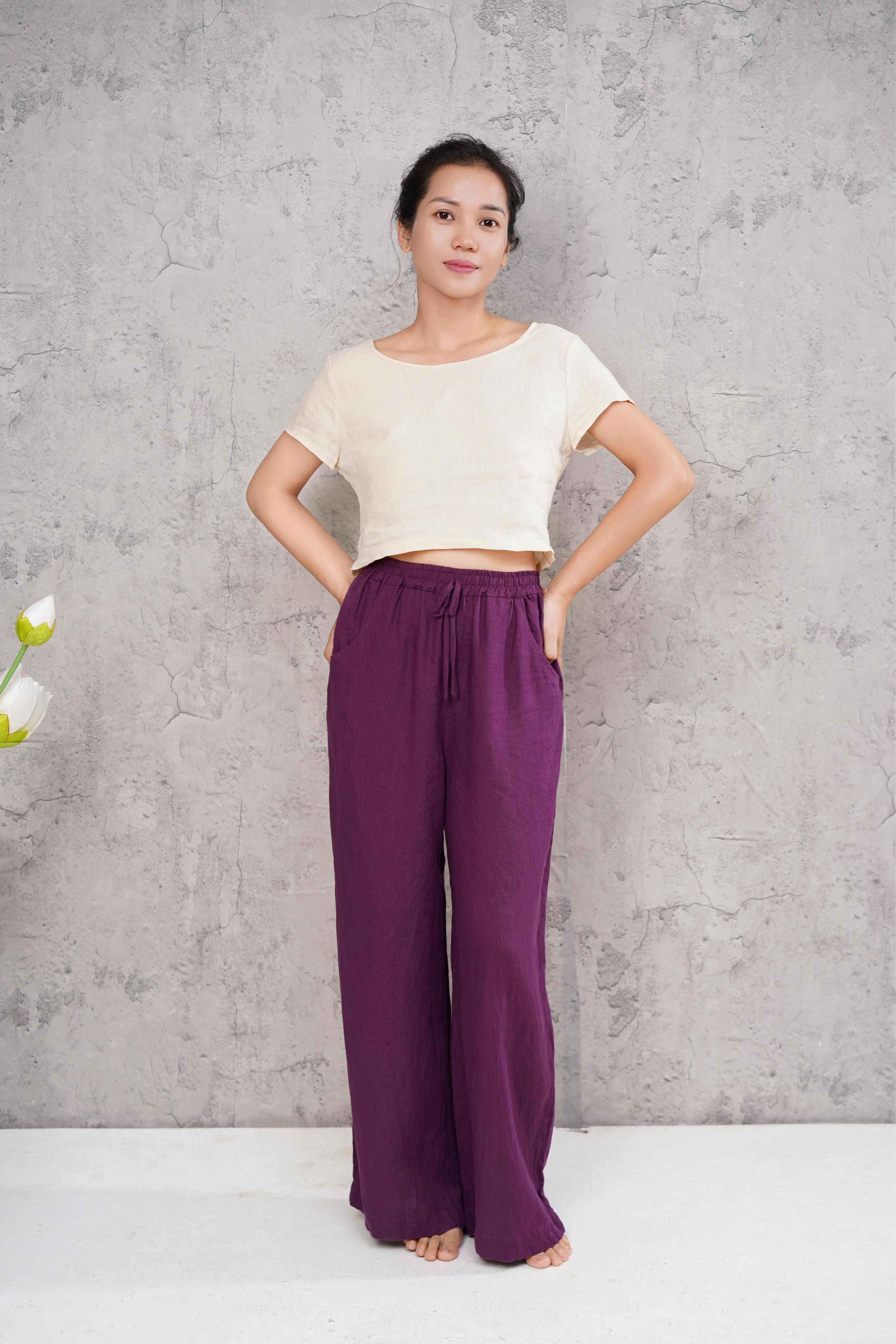 Woman wearing a white top and purple pants against a gray concrete wall.