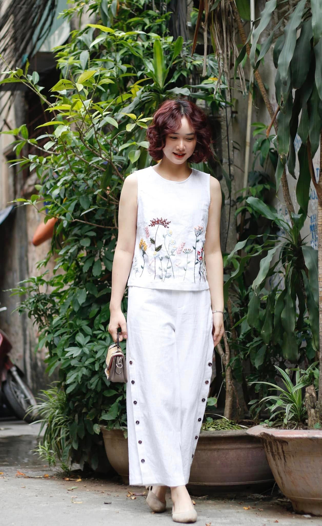 Woman in a white sleeveless top and pants standing among green plants.