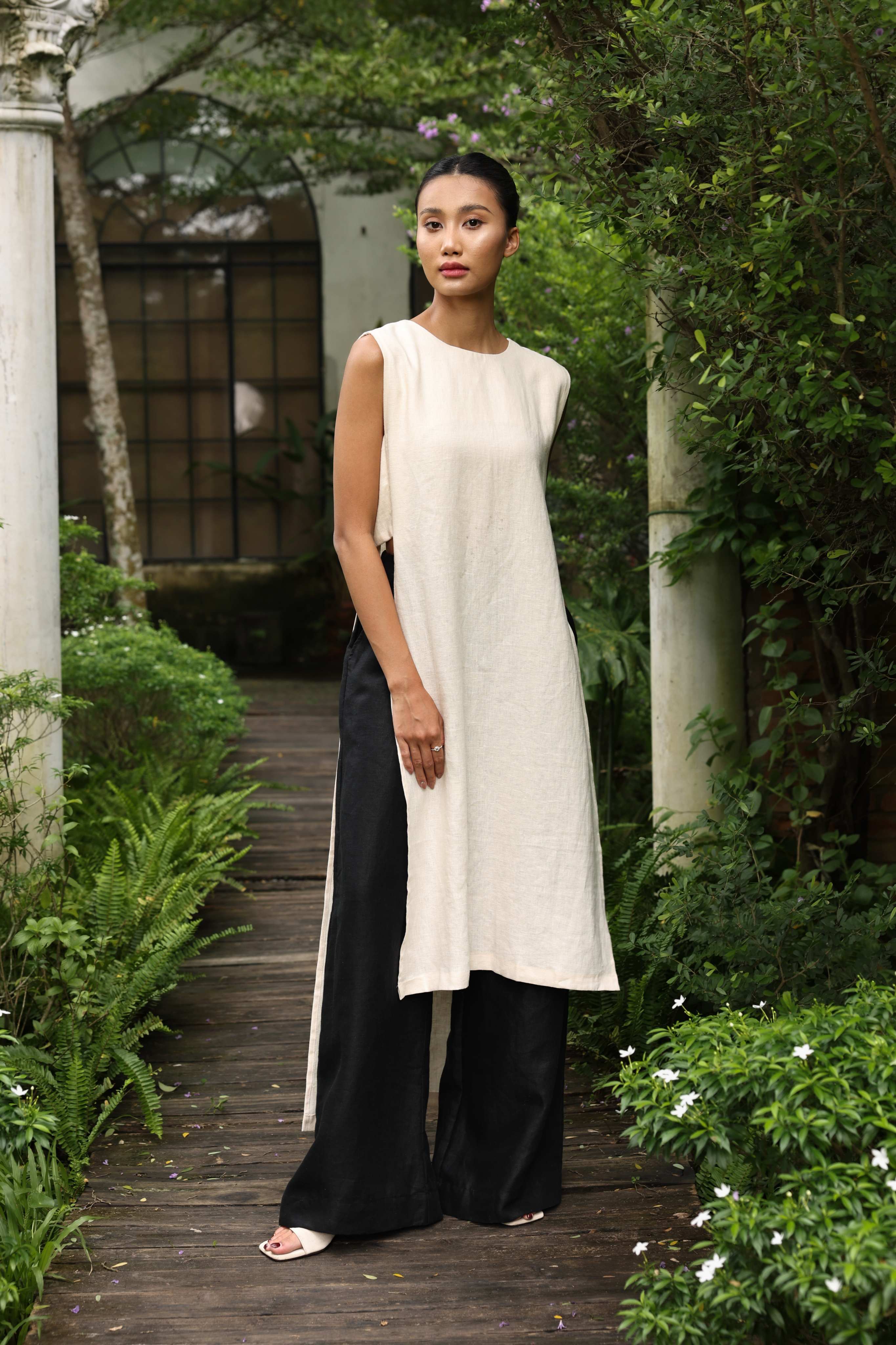 Woman wearing a white sleeveless top and black pants standing in a garden setting.