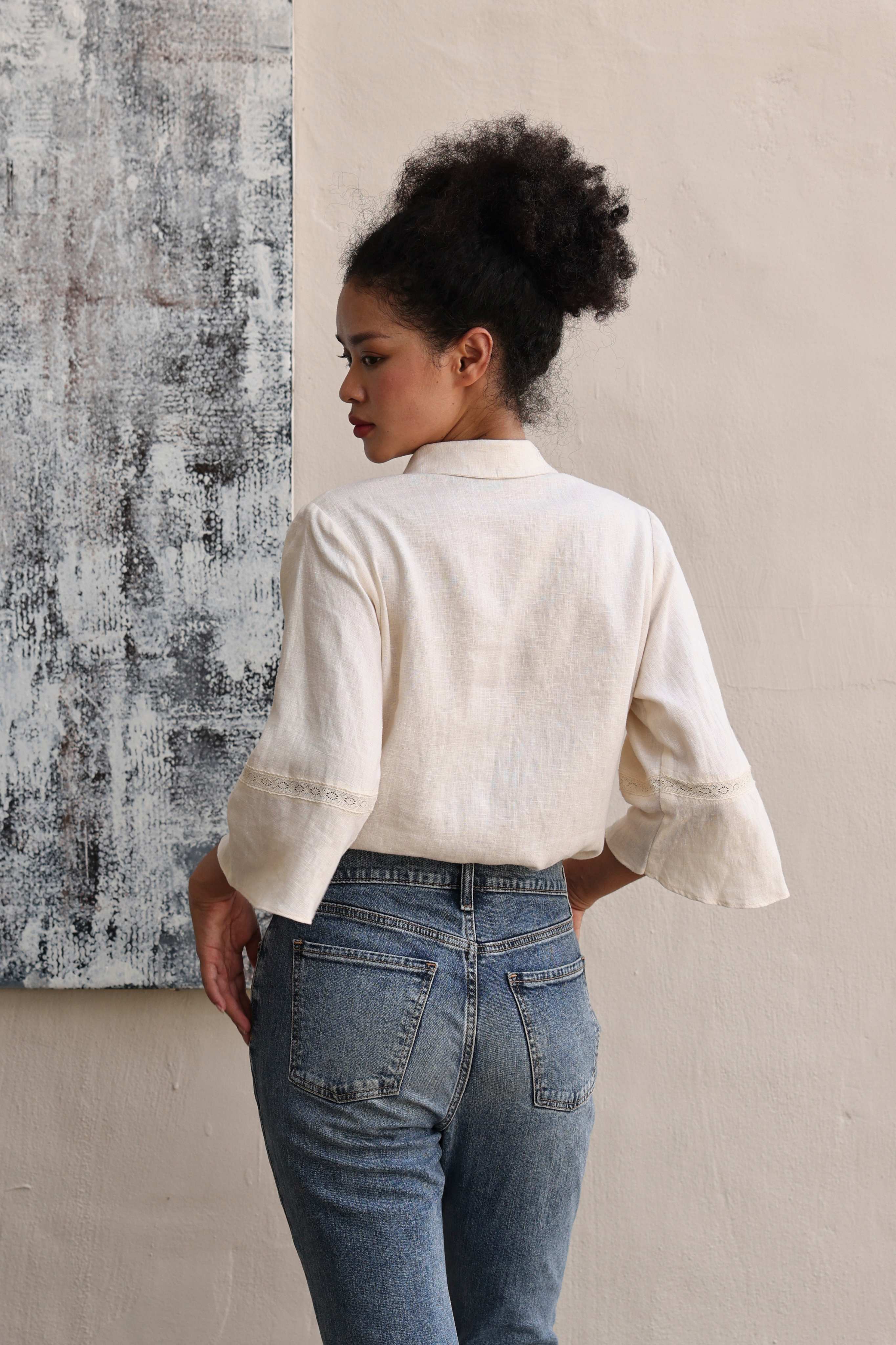 Woman wearing a beige blouse and blue jeans standing against a textured wall.