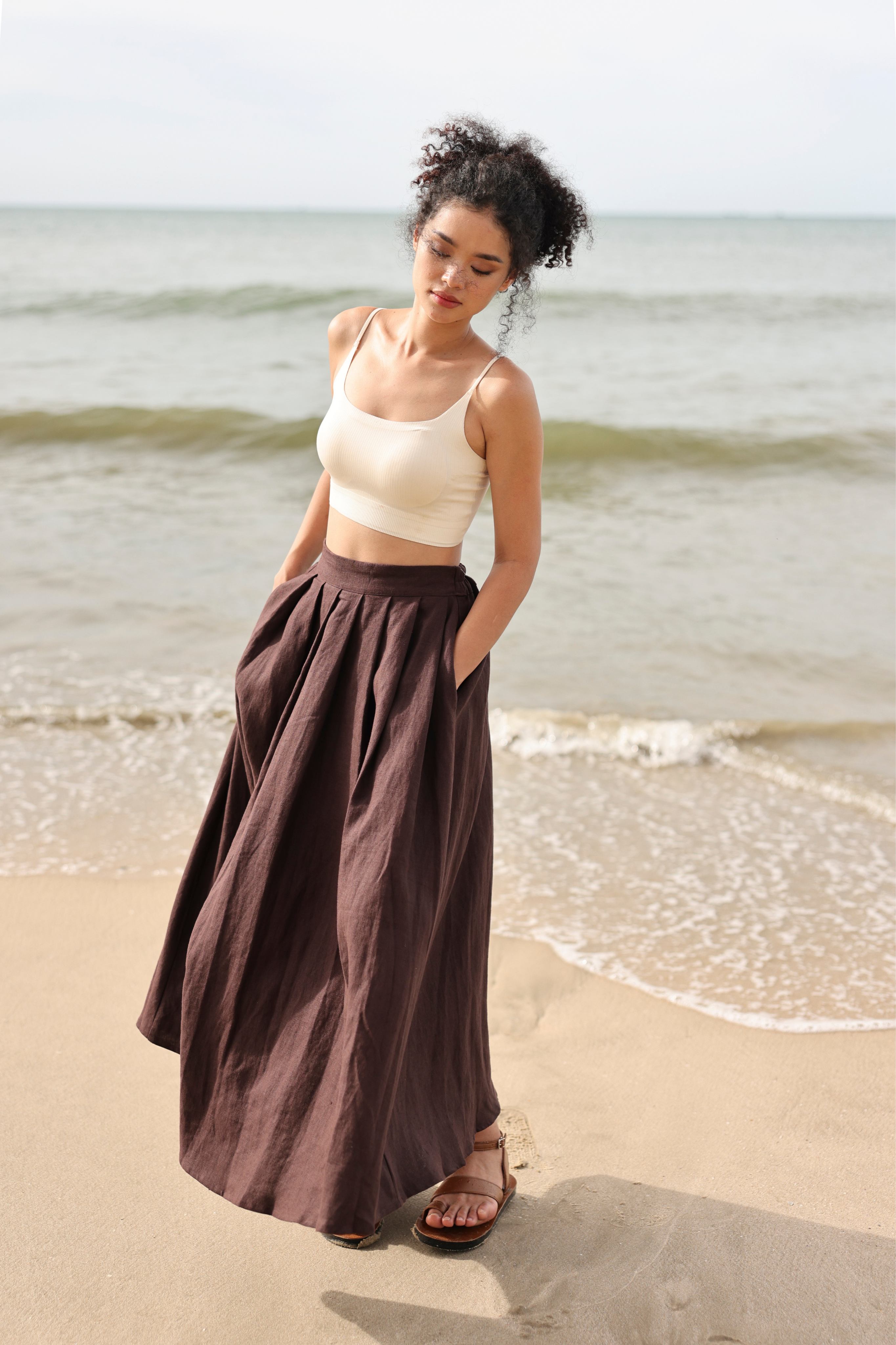 Woman standing on a beach wearing a white top and brown skirt.
