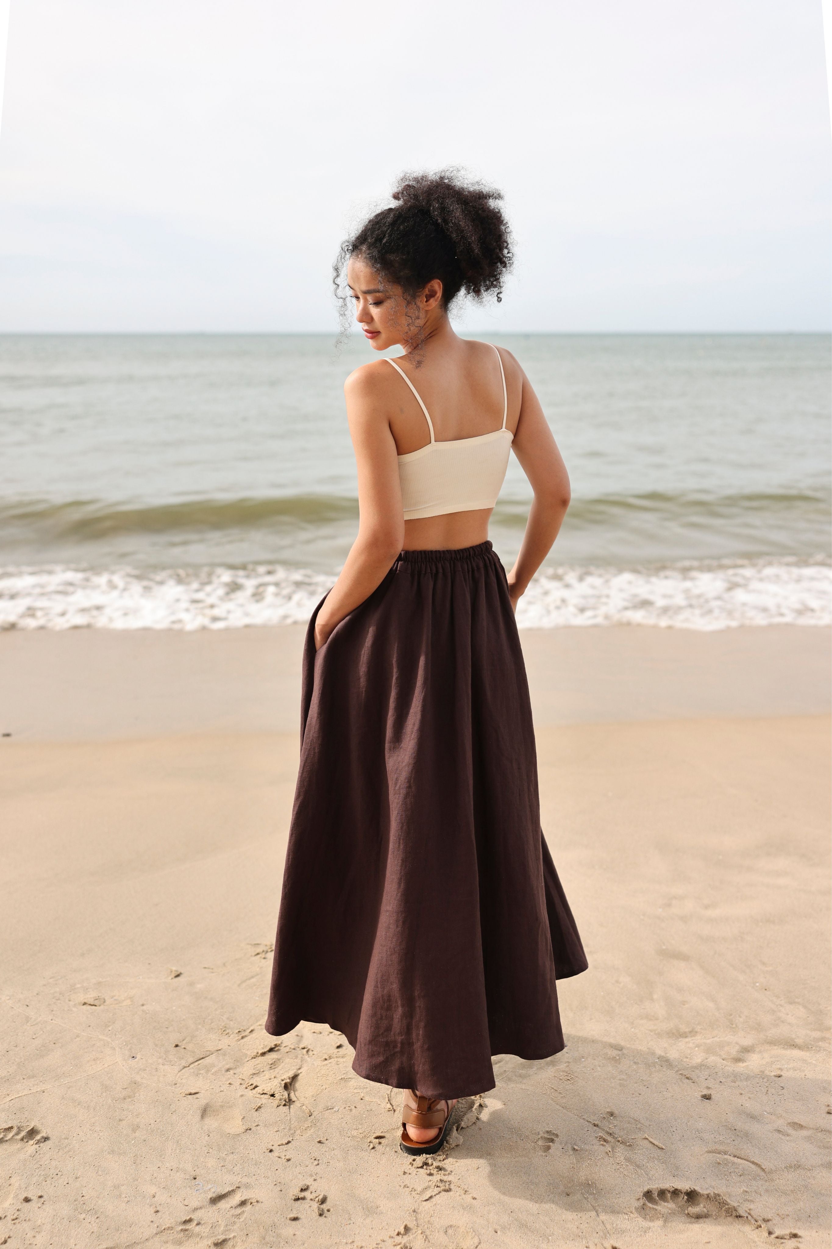 Woman standing on a beach wearing a beige top and brown skirt.