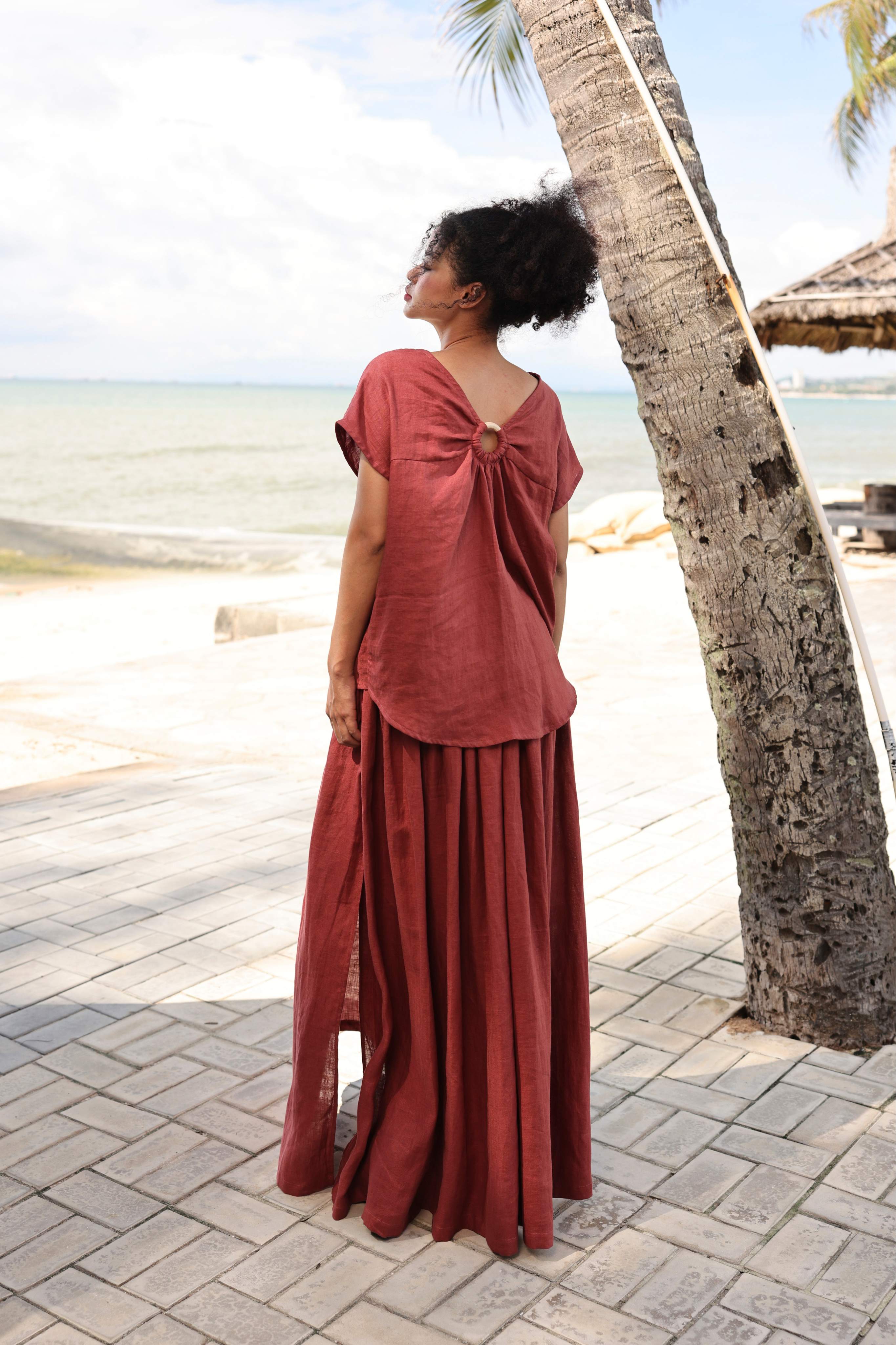 Woman in a rust-colored skirt standing by a palm tree by the beach.