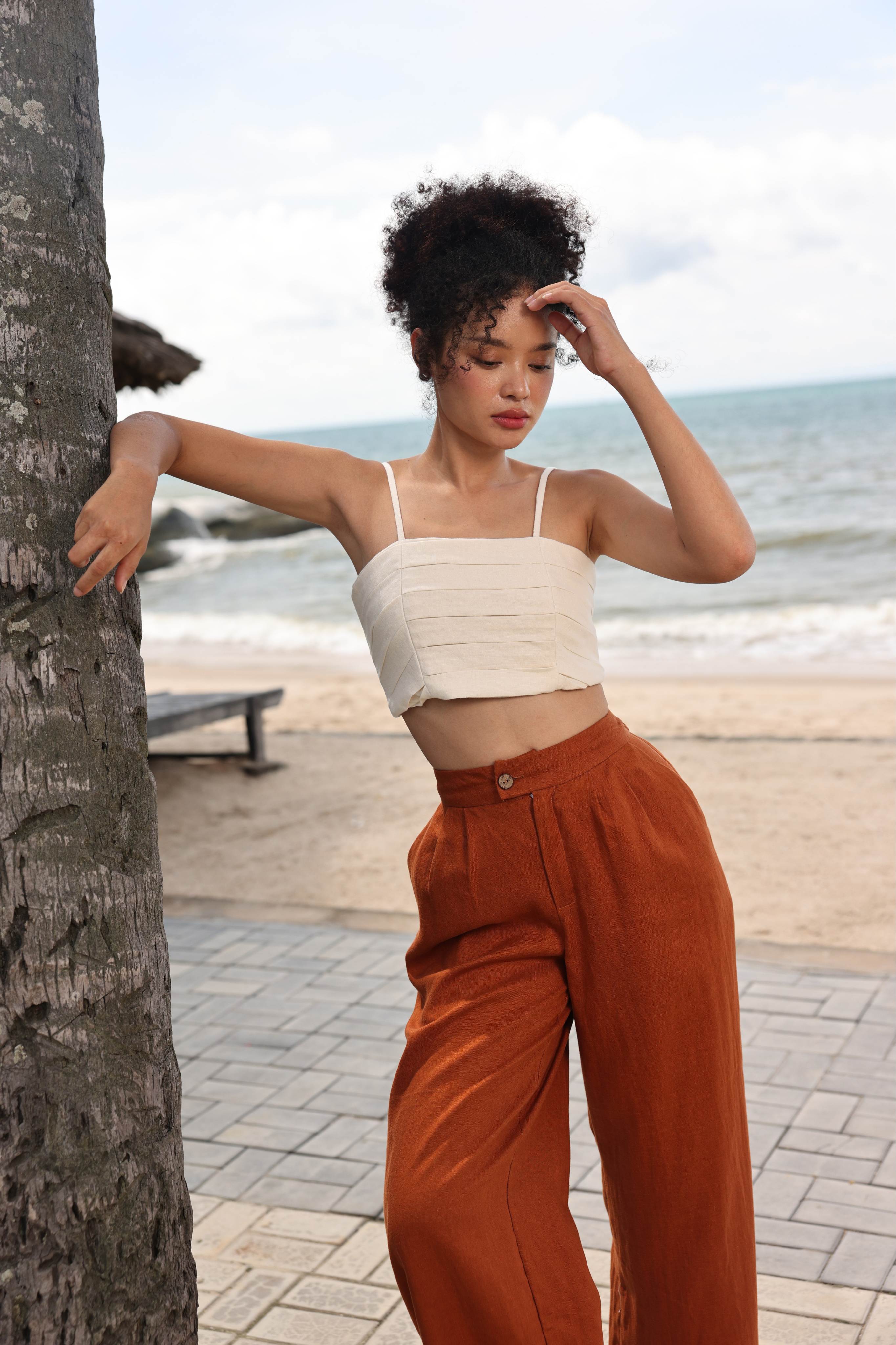 Woman in a top and rust-colored pants standing by a palm tree with a beach in the background