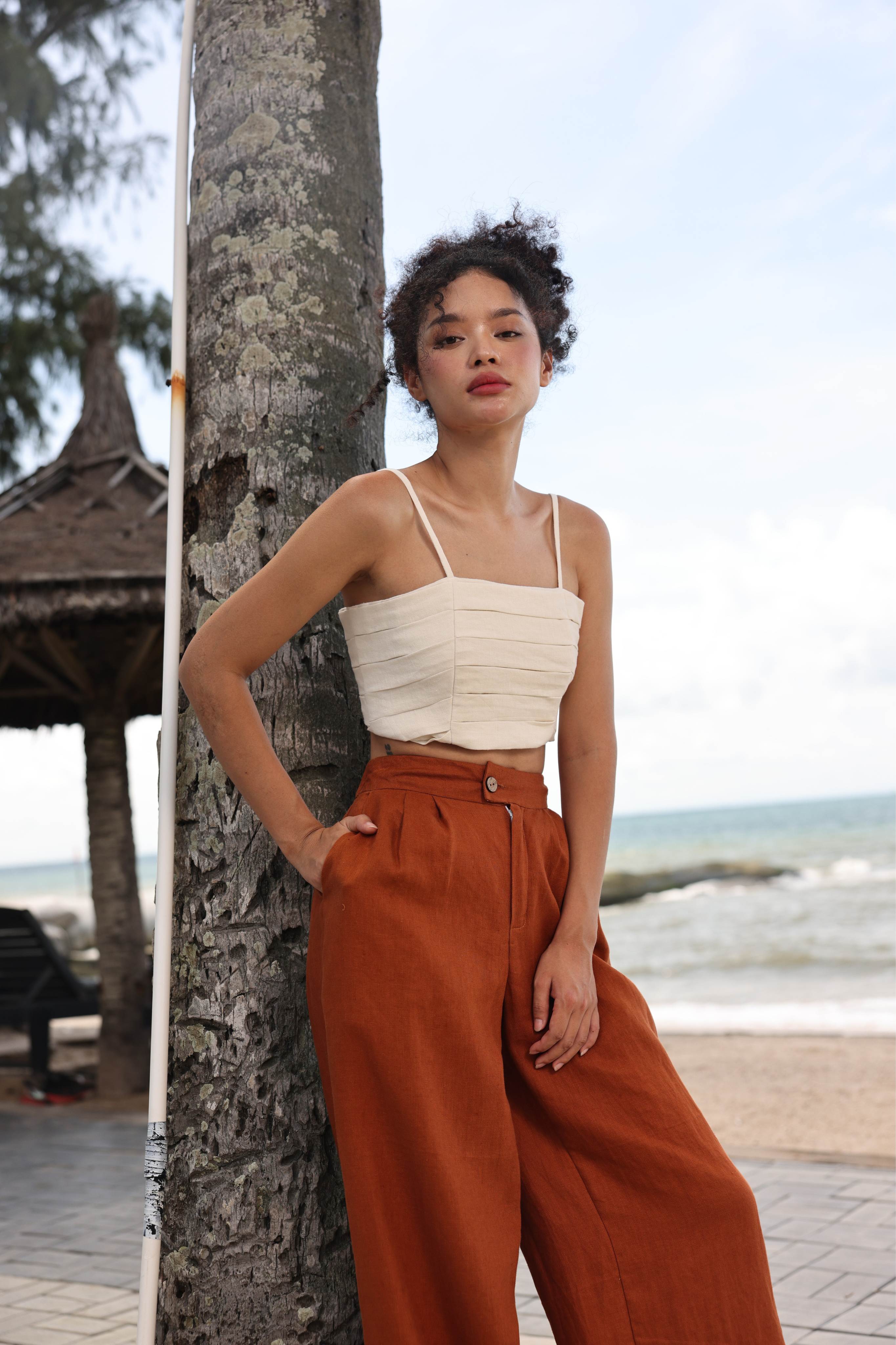 Woman in a beige top and rust-colored pants leaning against a tree by the beach.