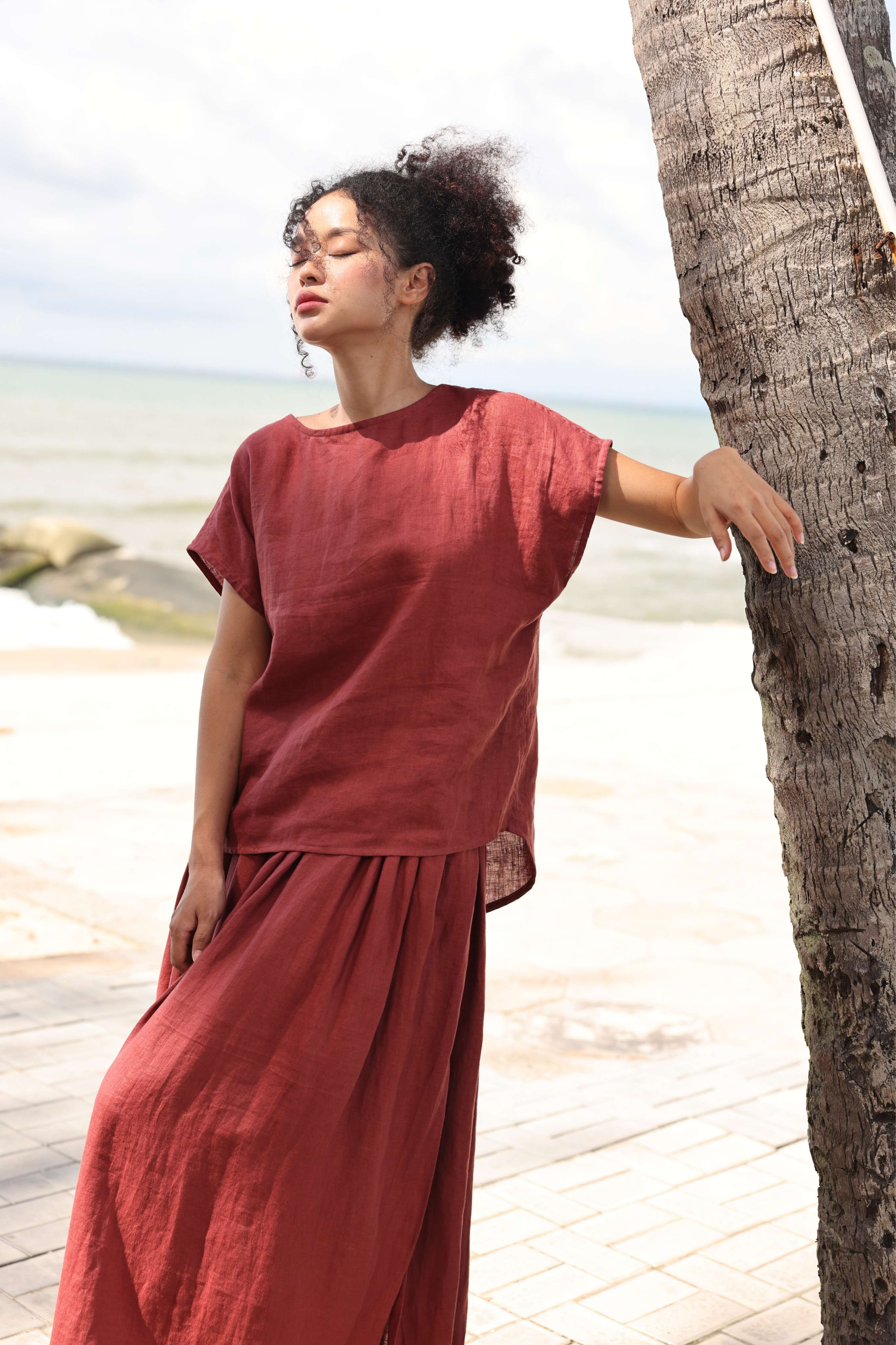 Woman in a red outfit leaning against a palm tree by the beach.