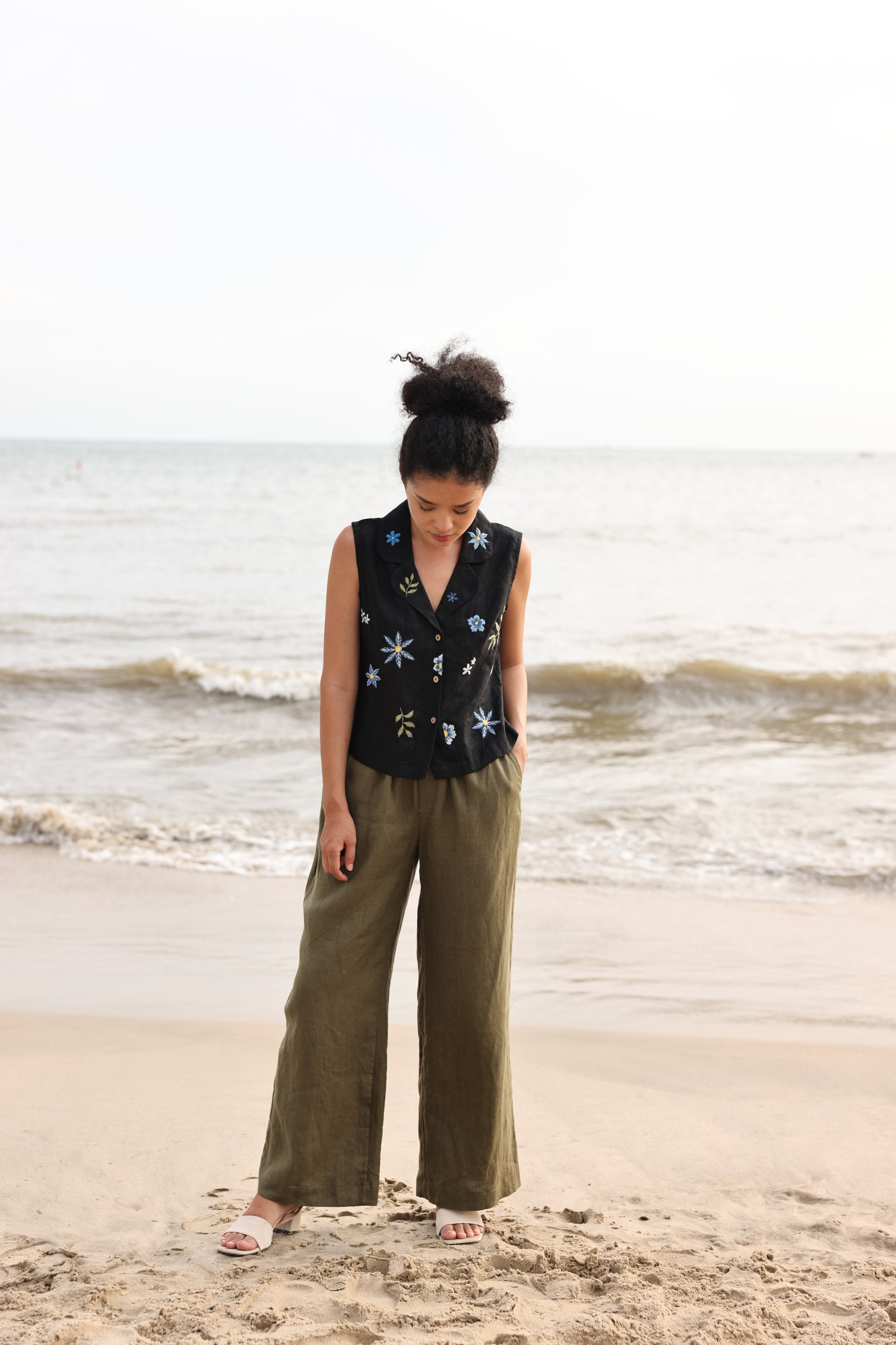 Woman standing on a beach wearing a sleeveless top with floral patterns and wide-leg pants.