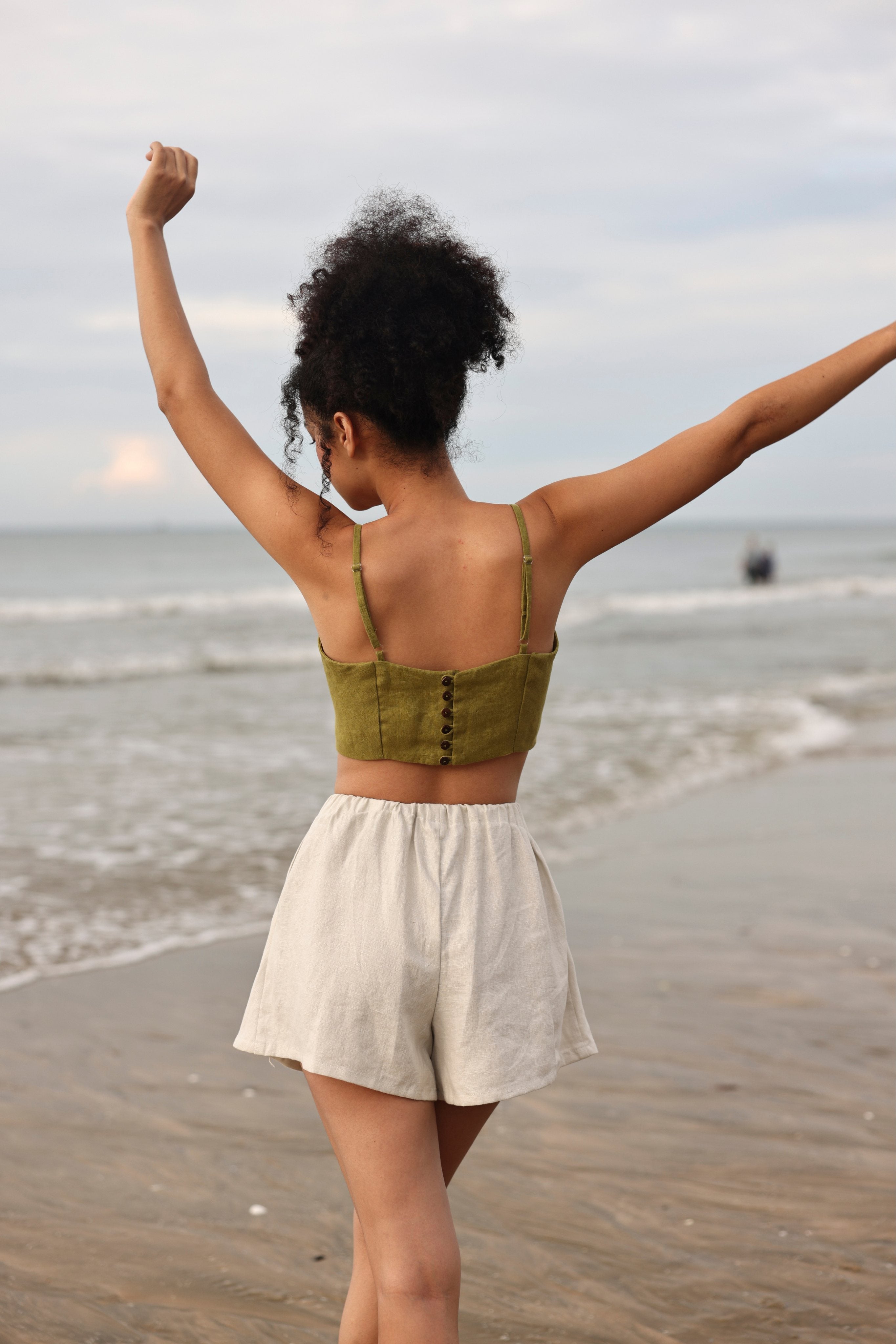 Woman standing on a beach with arms raised, wearing a green top and beige shorts.