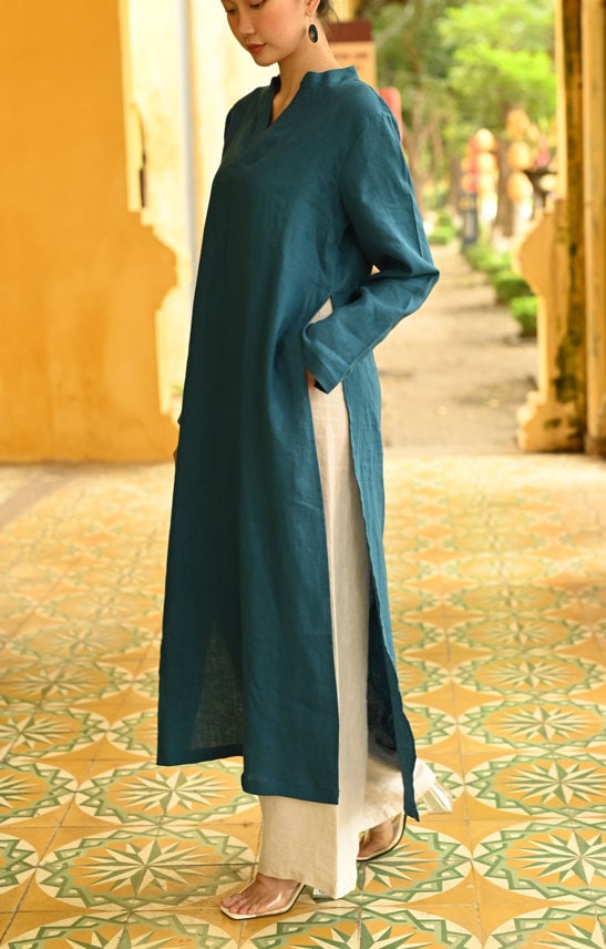 Person wearing a teal ao dai dress standing on a patterned floor with greenery in the background