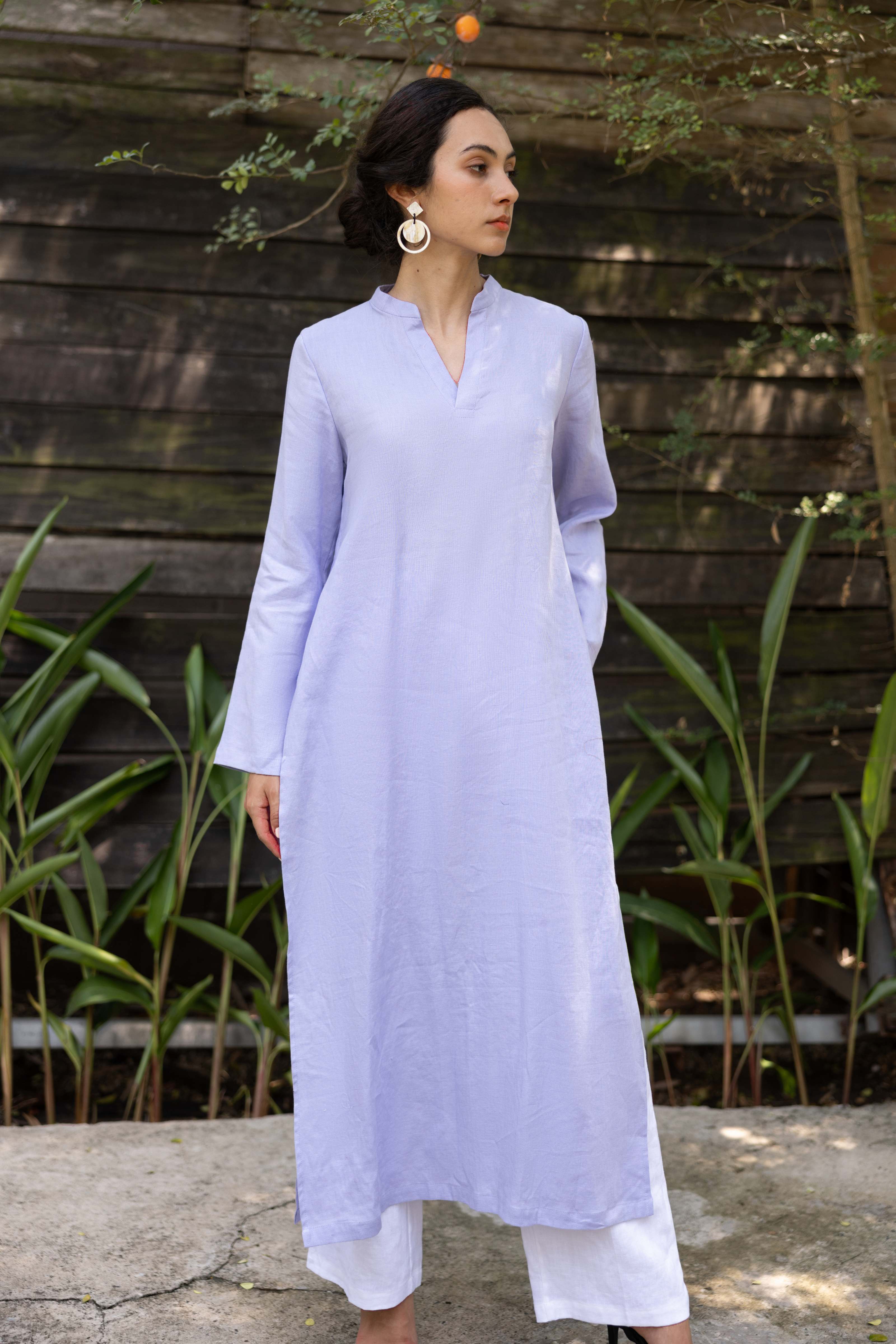Woman wearing a light purple dress standing outdoors with a wooden wall and plants in the background
