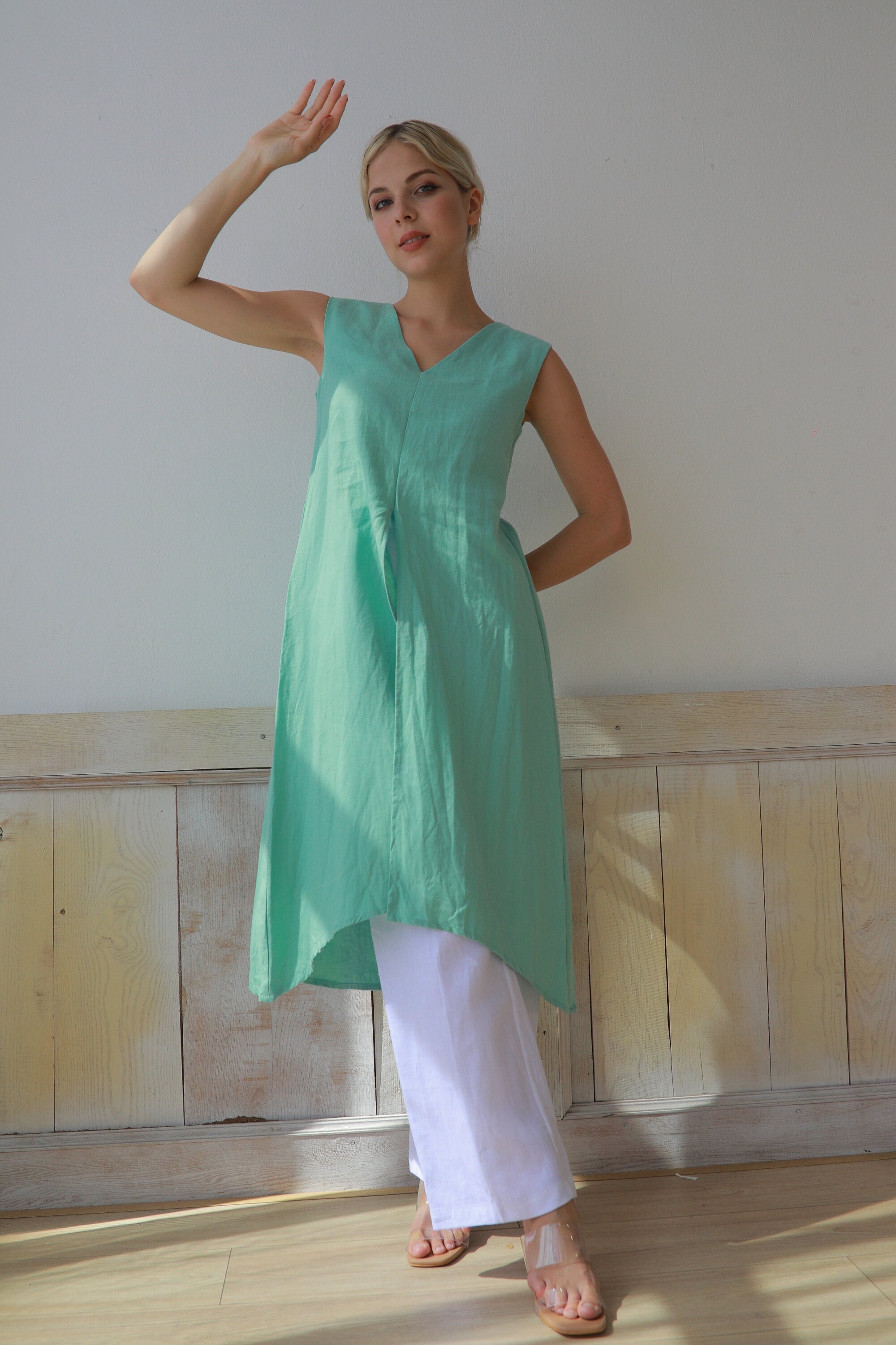 Woman wearing a green tunic with white pants against a plain wall.