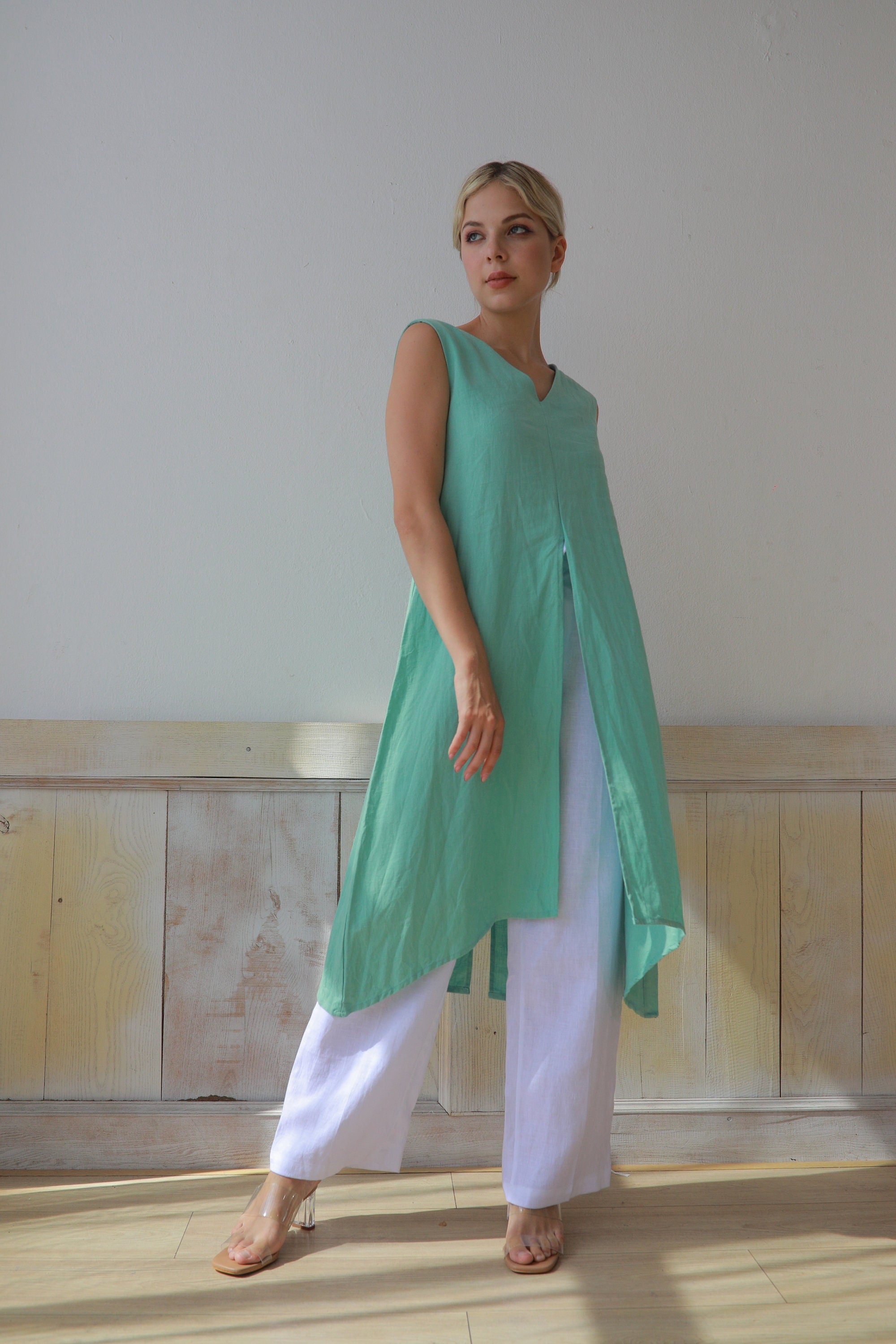 Woman wearing a green sleeveless top and white pants against a plain wall.