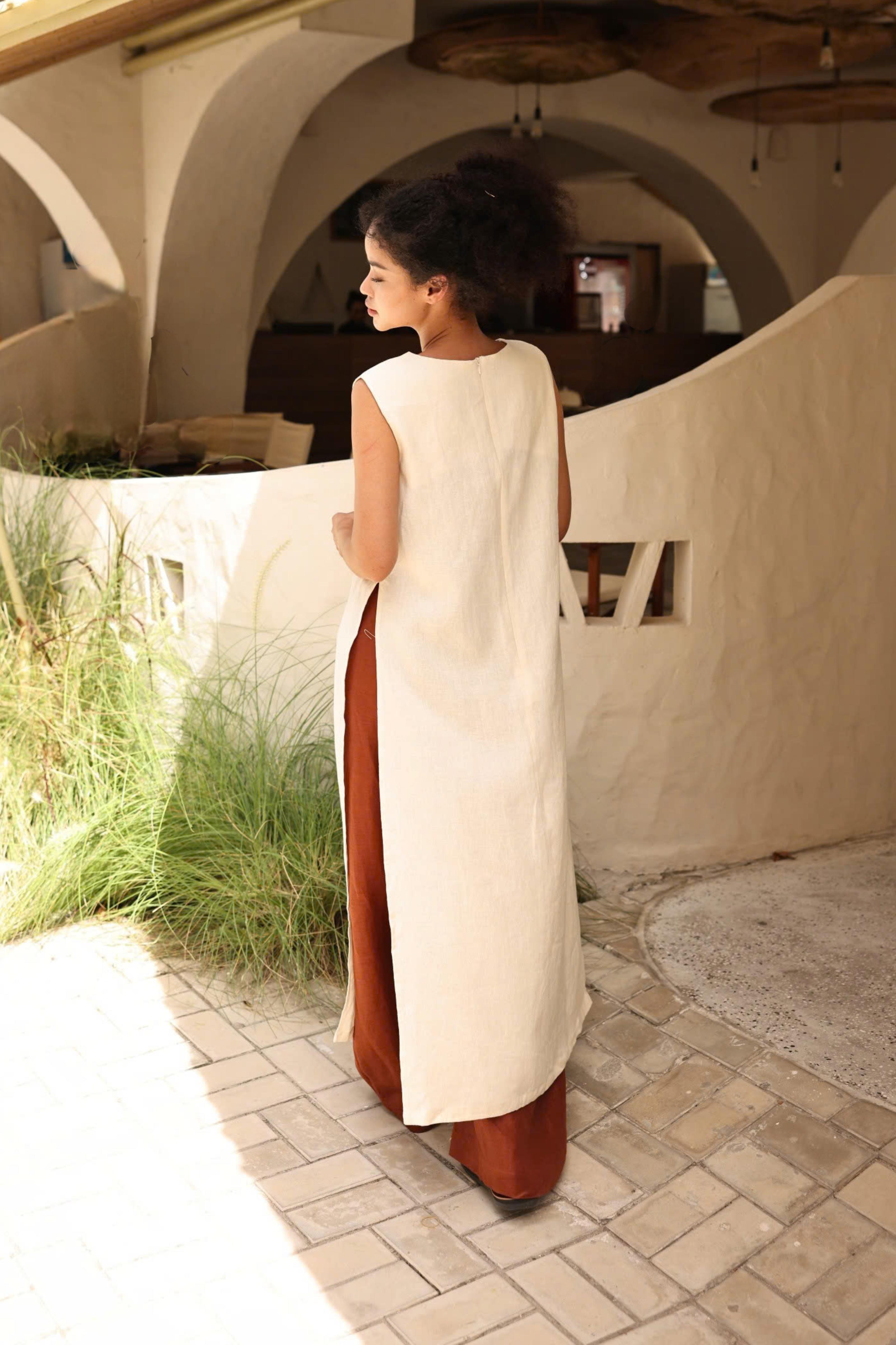 Woman in a long, light-colored dress standing on a stone path with greenery and architectural elements in the background.