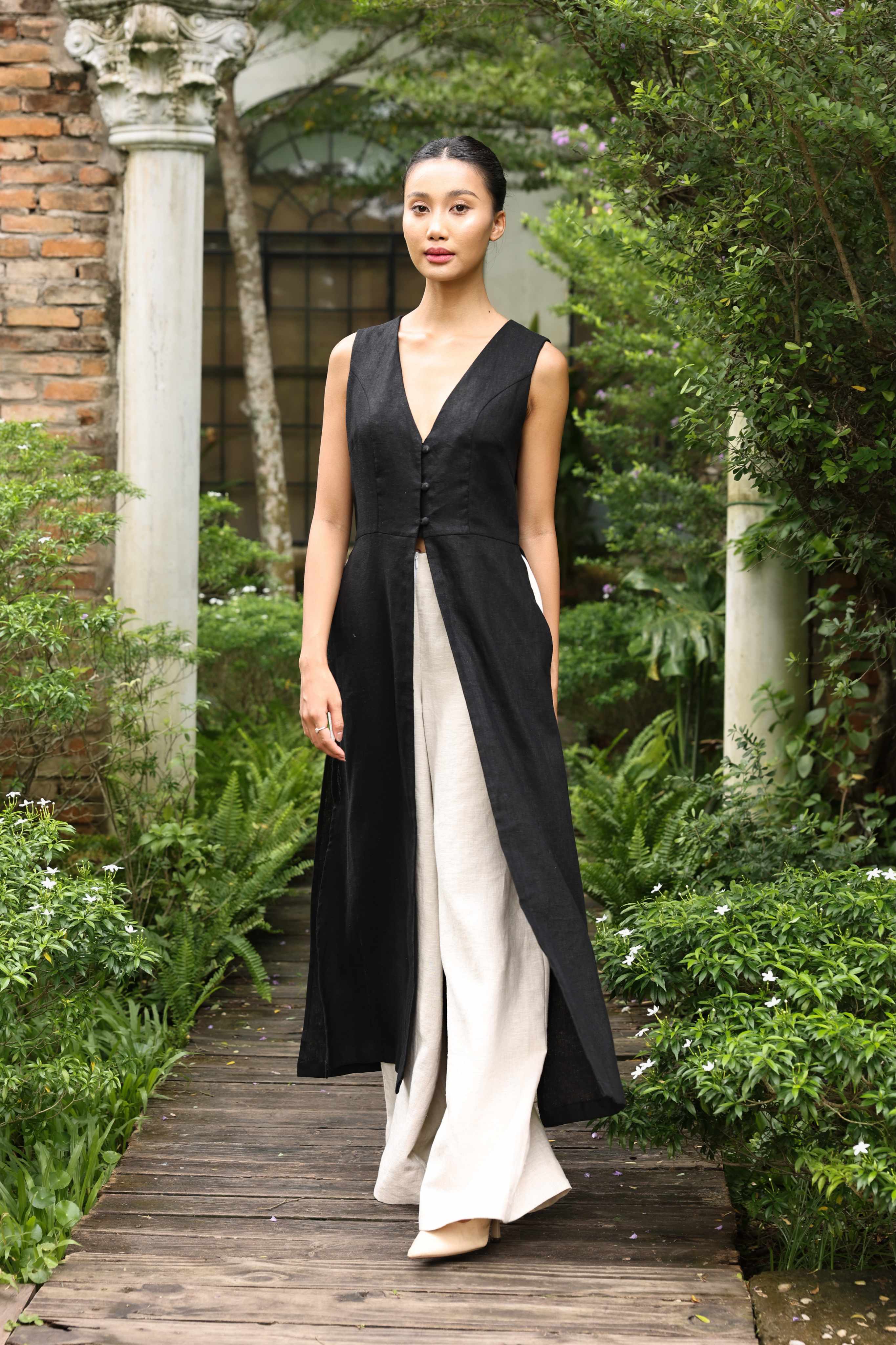 Woman wearing a black tunic and white pants standing on a wooden path in a garden.