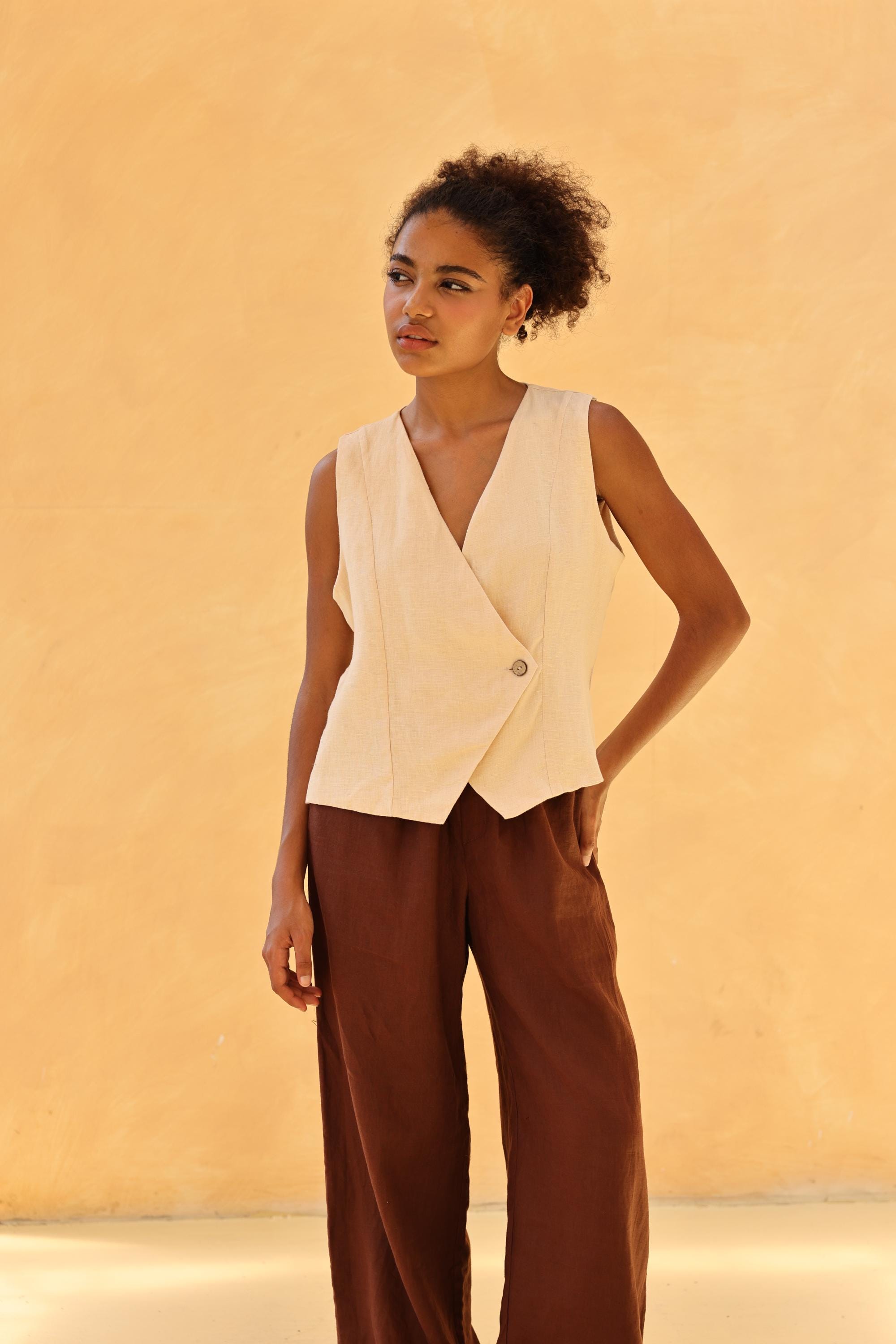 Woman wearing a beige sleeveless blazer over brown pants against a beige background