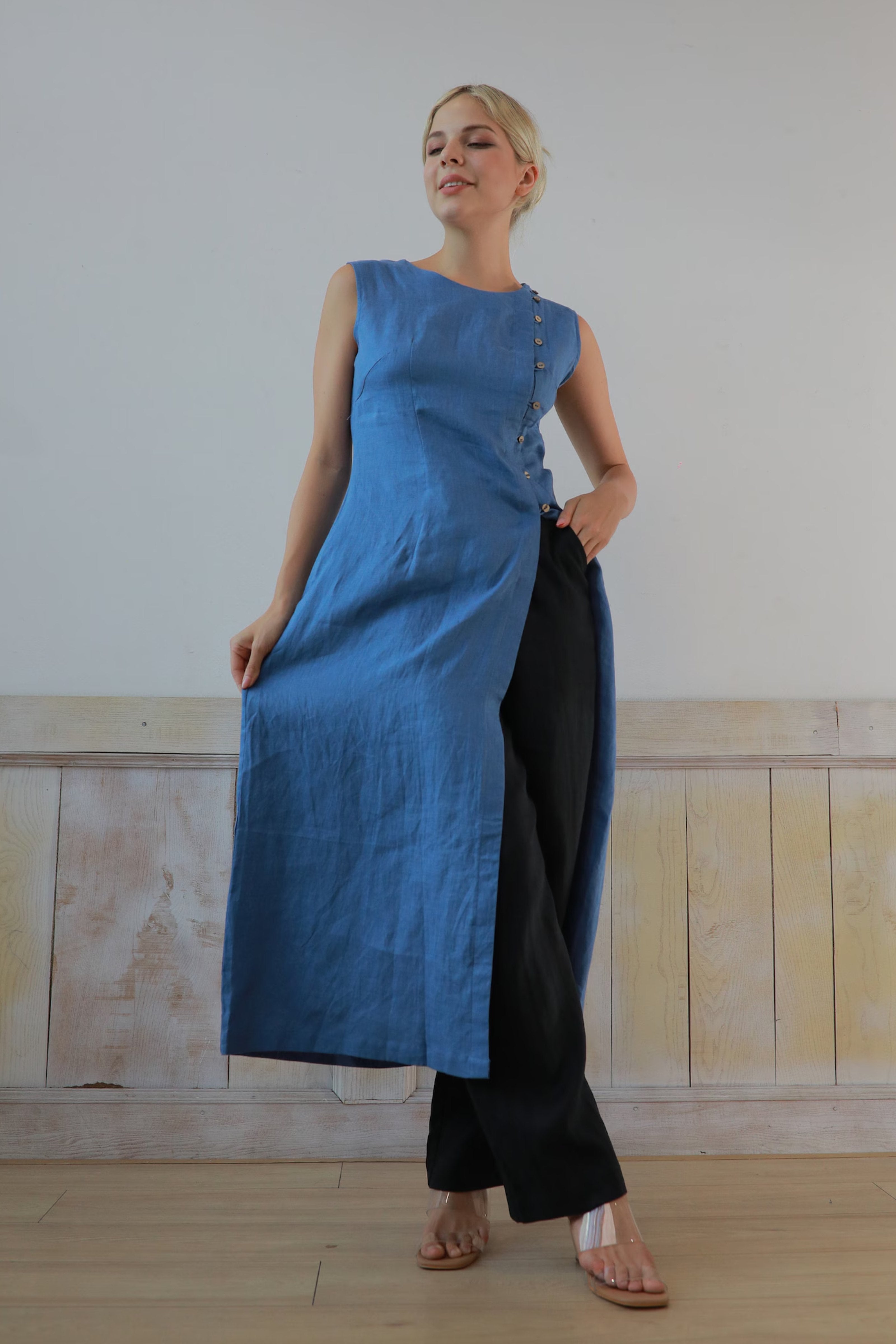 Woman wearing a blue sleeveless top and black pants against a plain wall.