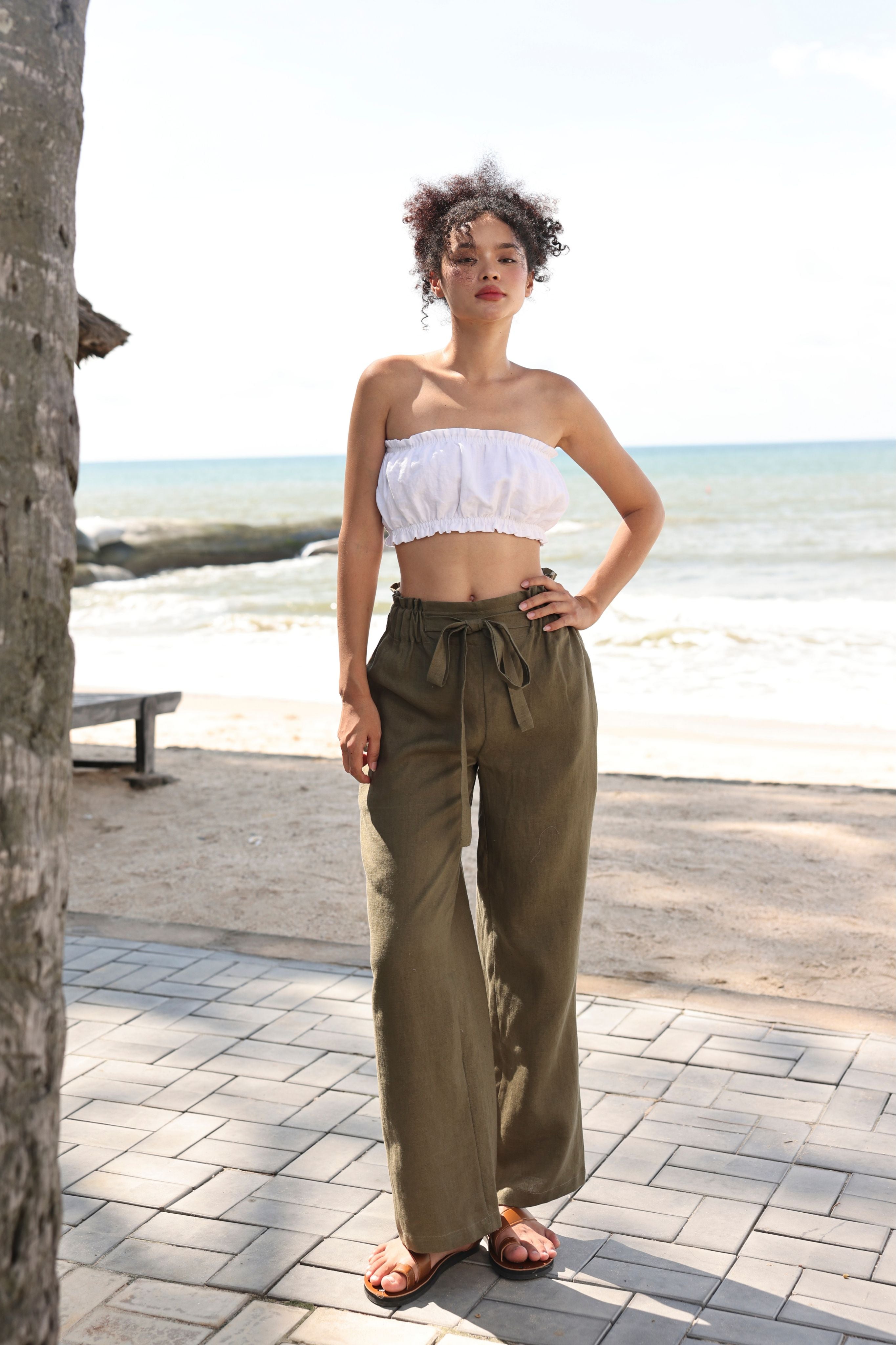 Woman in white top and olive green pants standing on a beach path with ocean view.