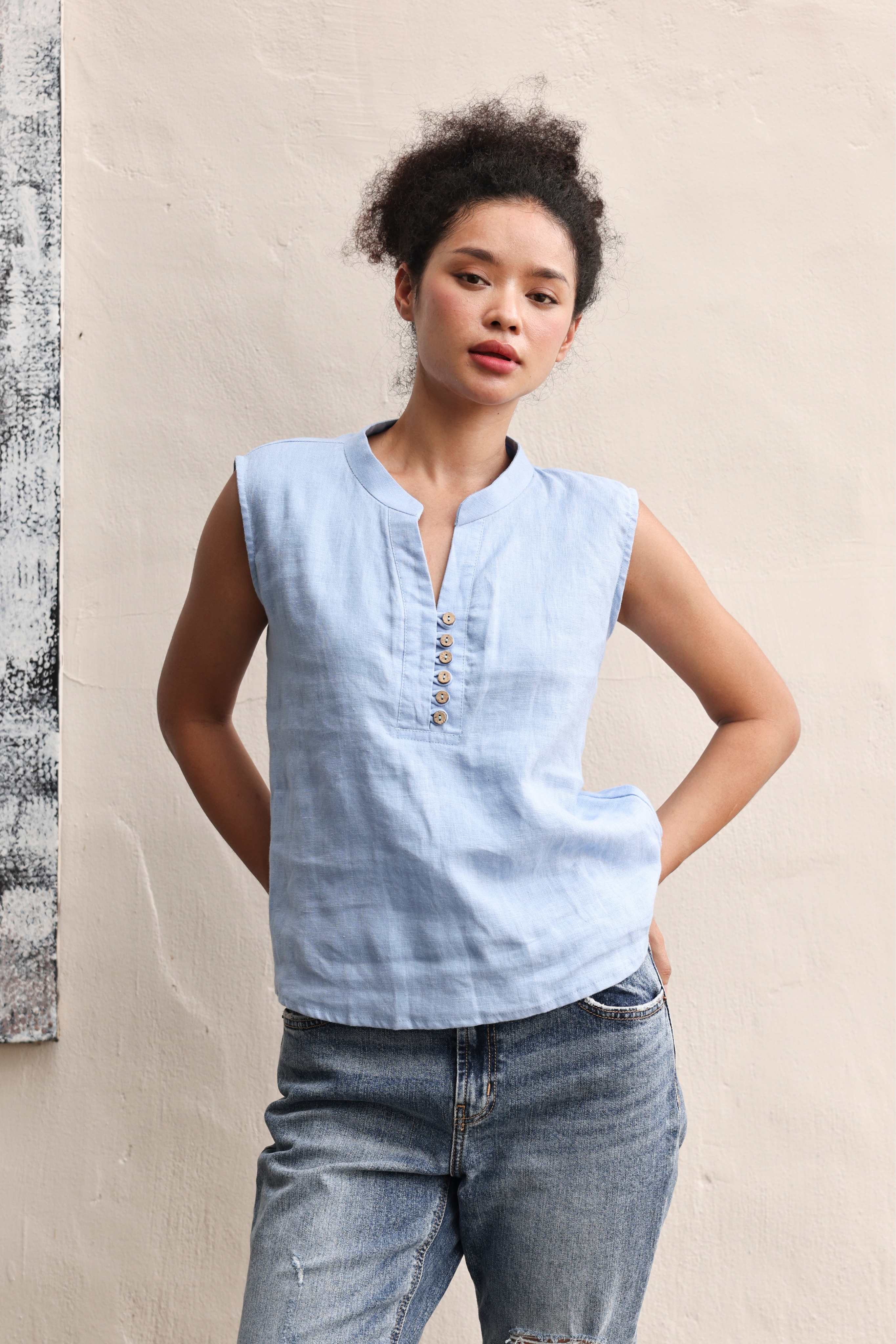 Woman wearing a light blue sleeveless shirt and jeans against a beige wall.