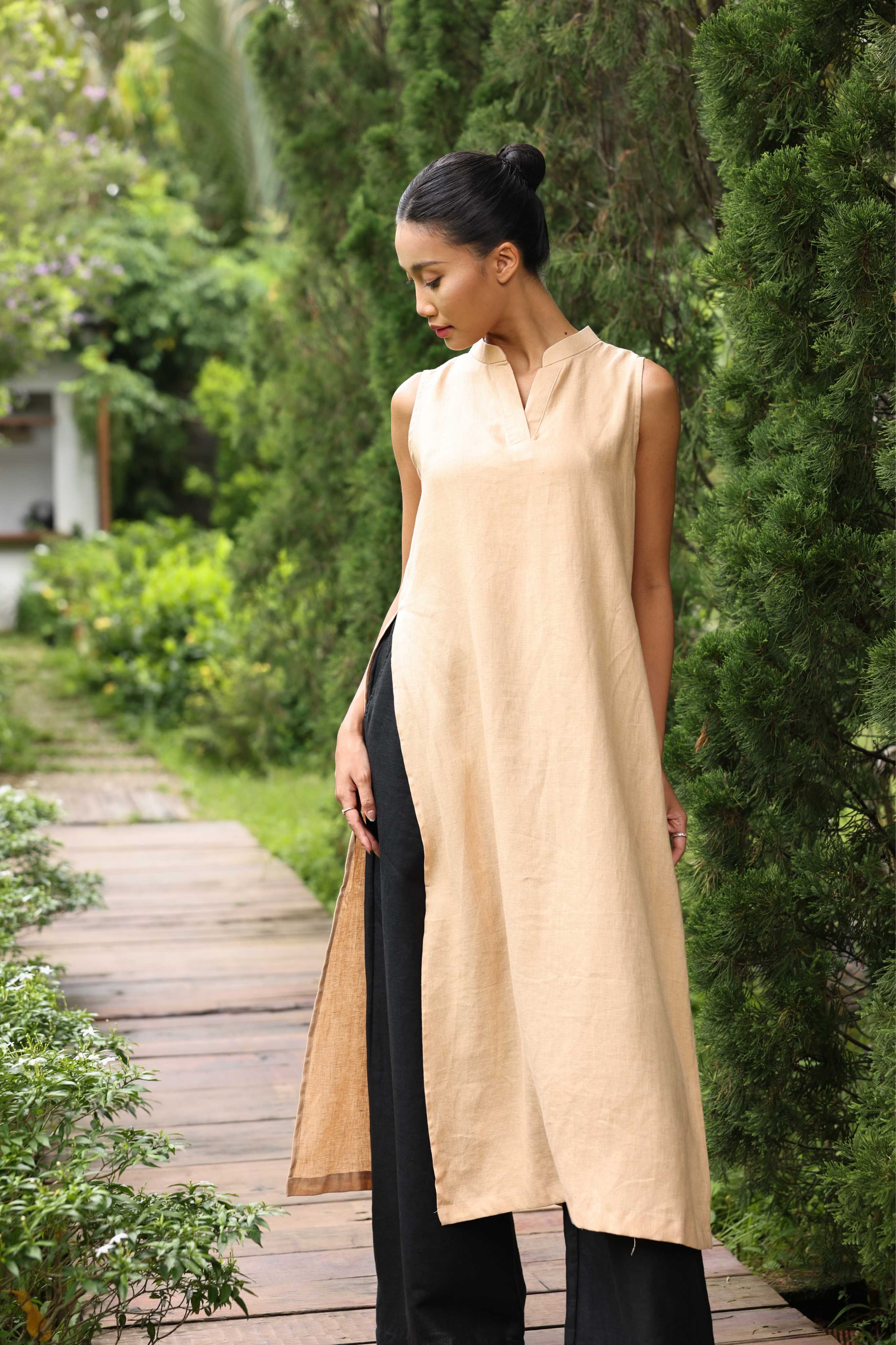 Woman wearing a beige sleeveless top and black pants walking on a wooden path in a garden.