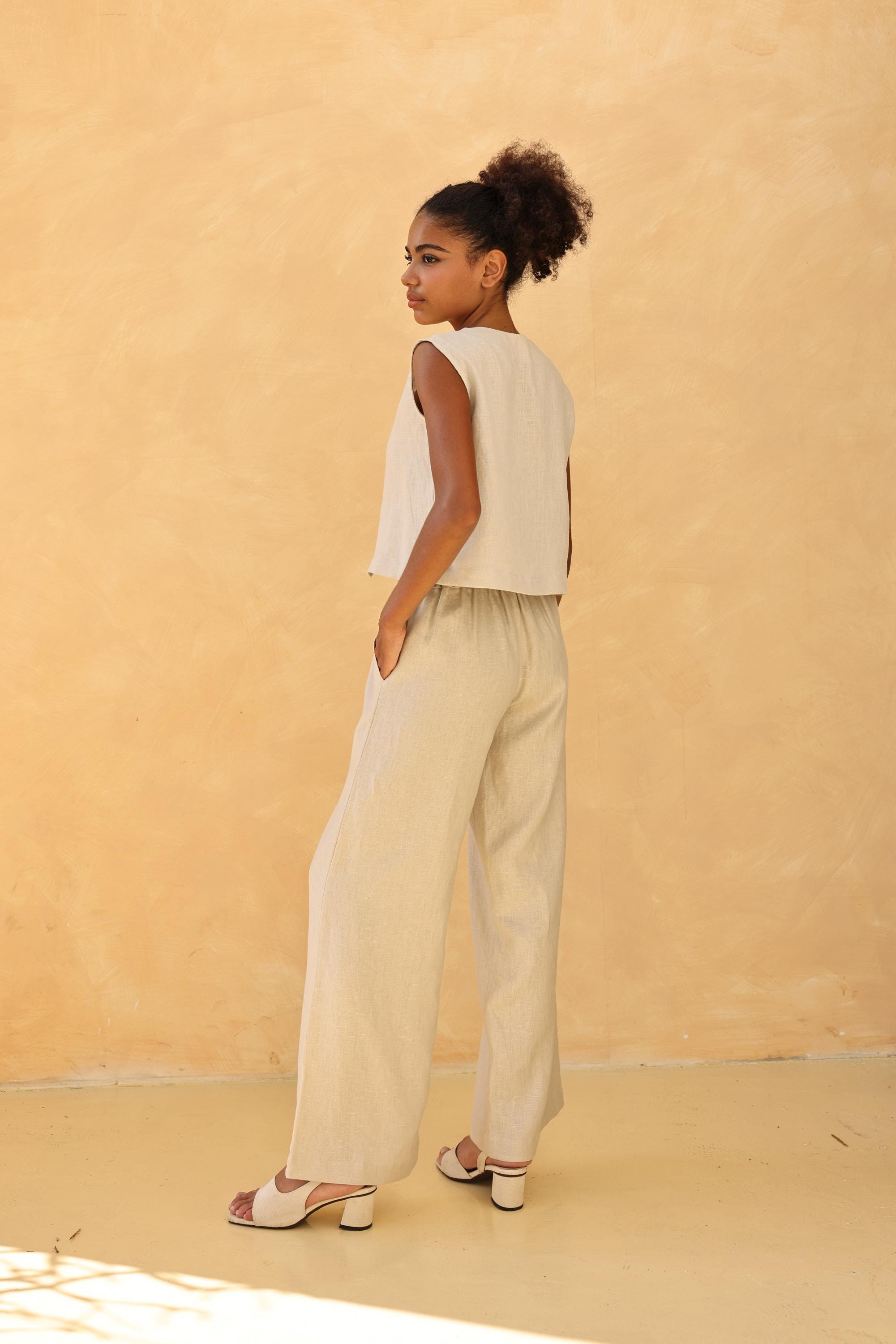 Woman wearing a sleeveless top and beige pants against a beige wall.