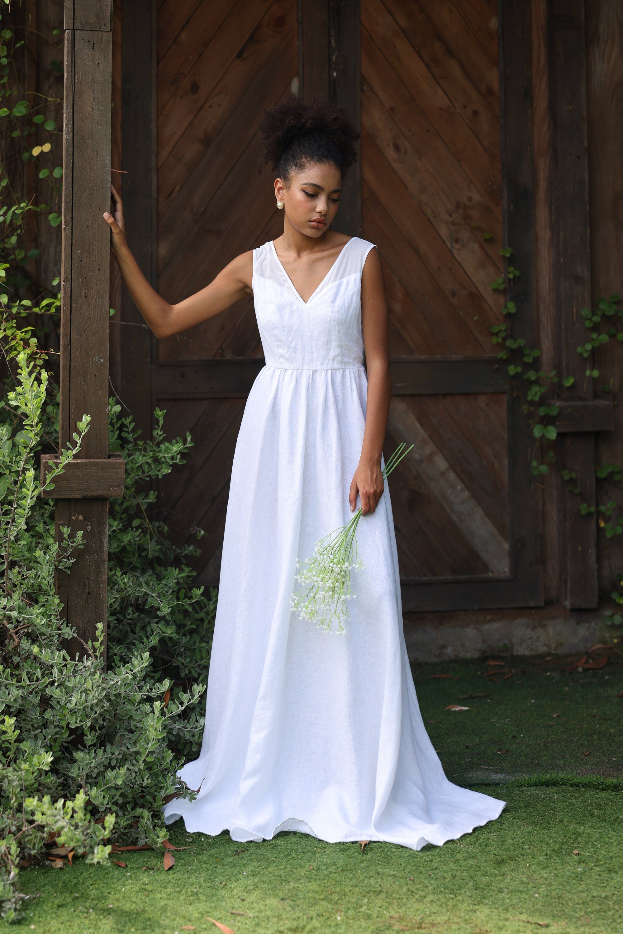 Woman in a white dress standing outdoors with greenery and a wooden wall in the background