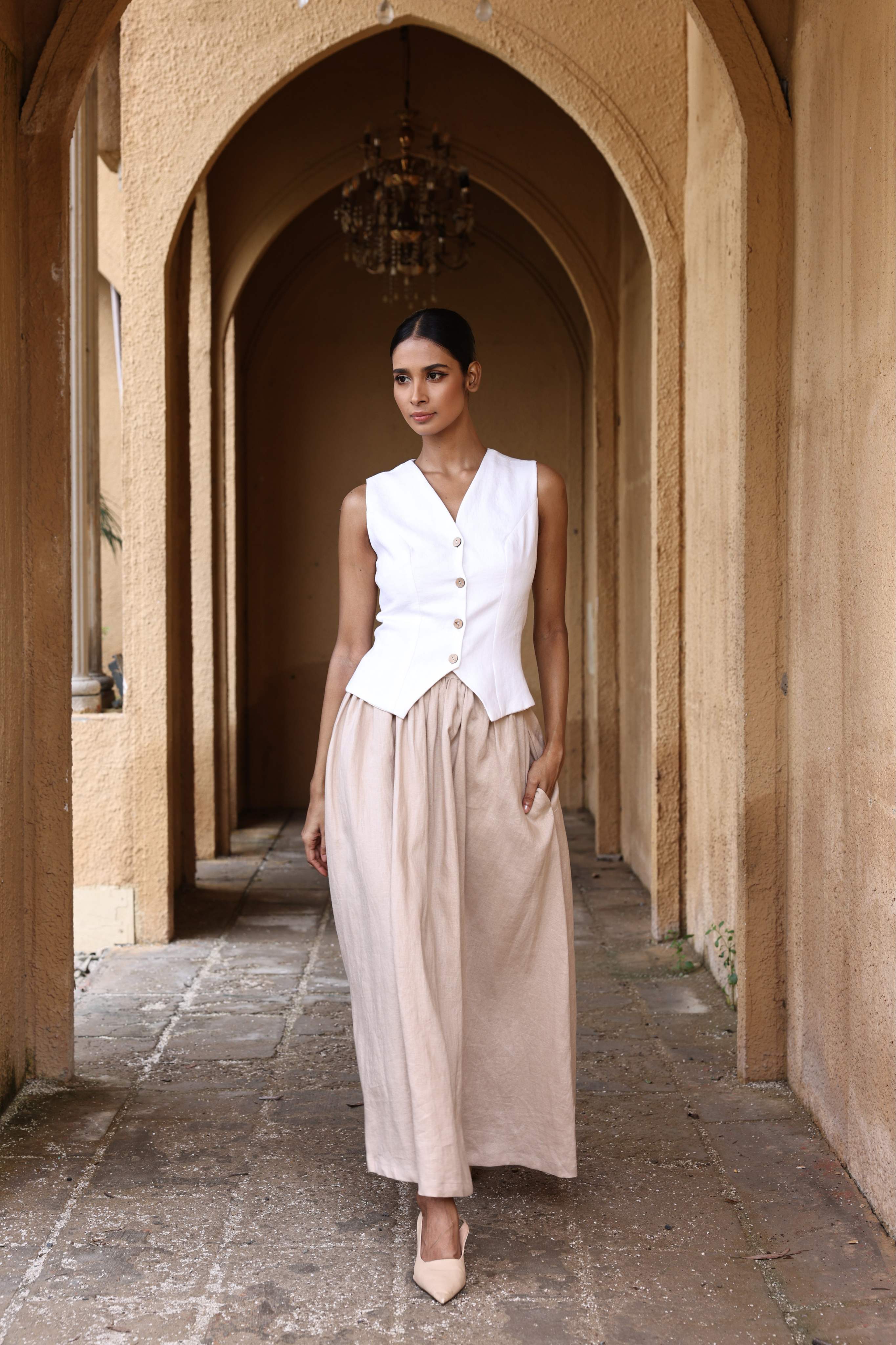 Woman in a white sleeveless top and beige skirt standing in an arched stone corridor.